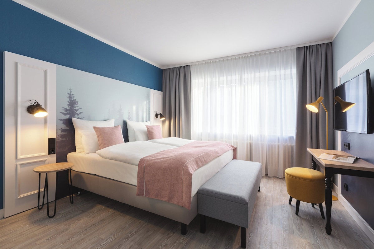 Quarto de hotel moderno com uma cama grande, mesas de cabeceira, uma escrivaninha e uma televisão.