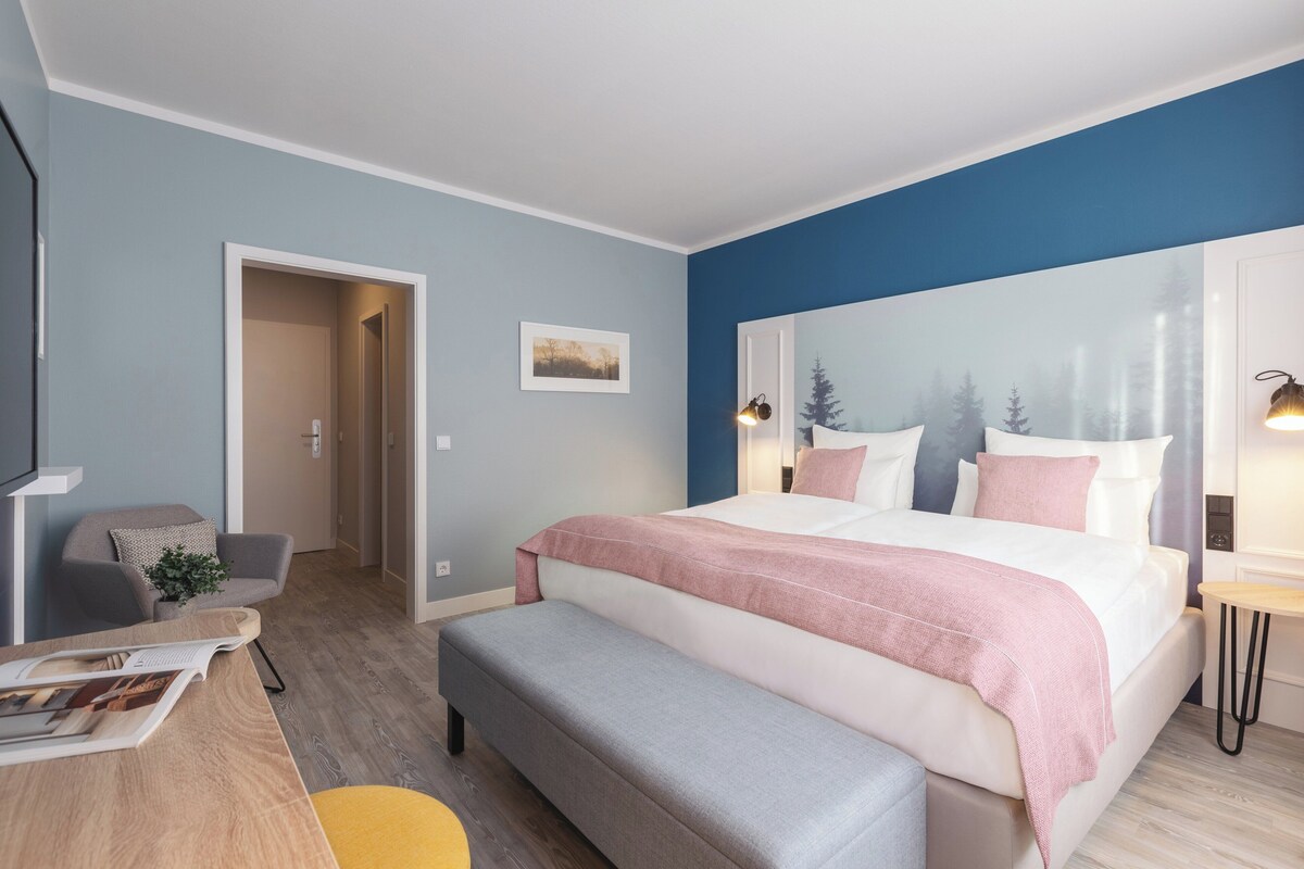 Quarto moderno com uma cama grande, uma escrivaninha com cadeira, um banco e uma luminária na parede.