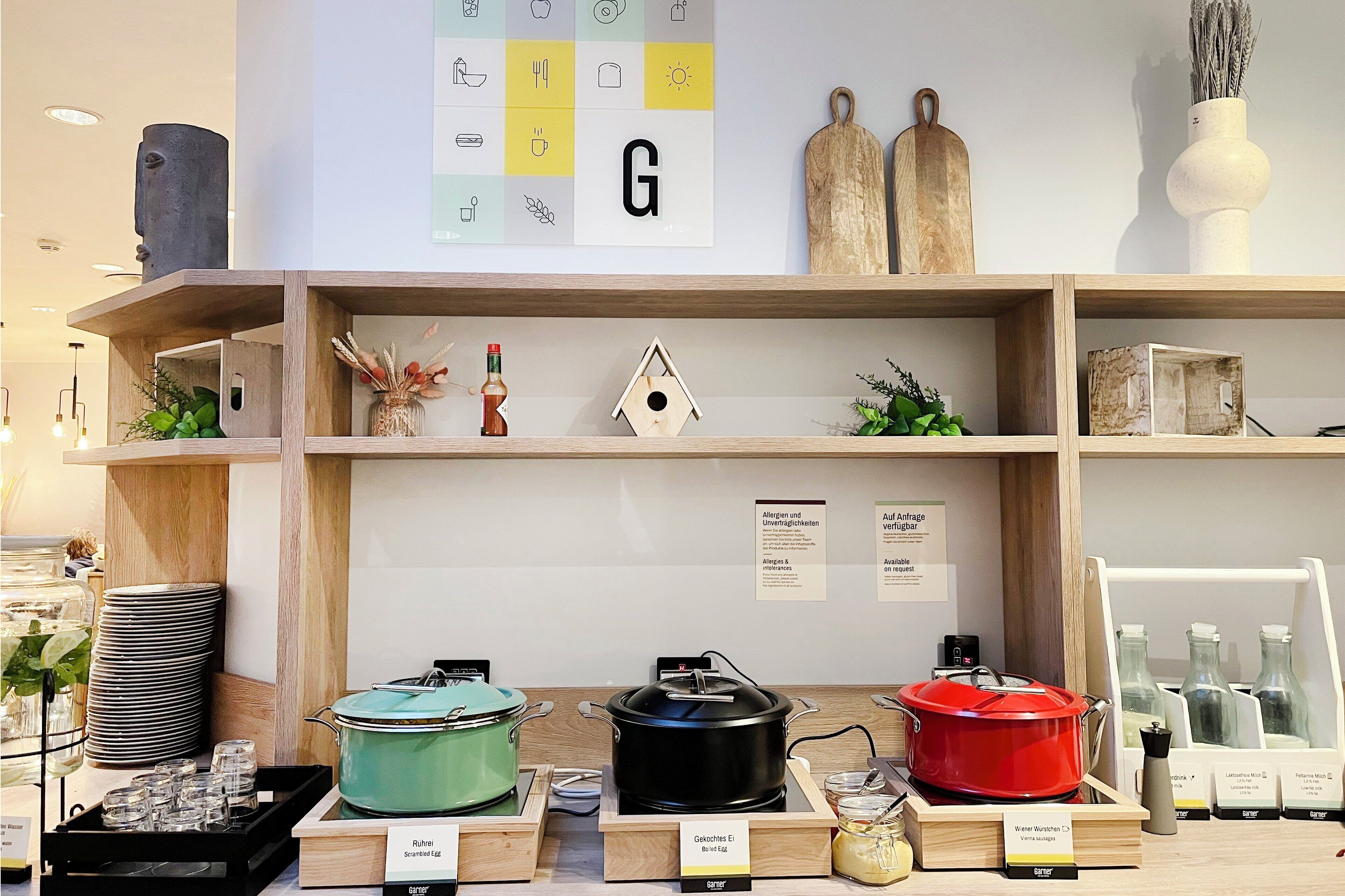 A kitchen area with wooden shelves displaying various kitchenware, including pots, pans, and a kettle. There are also stacks of plates and a sign indicating allergen information.