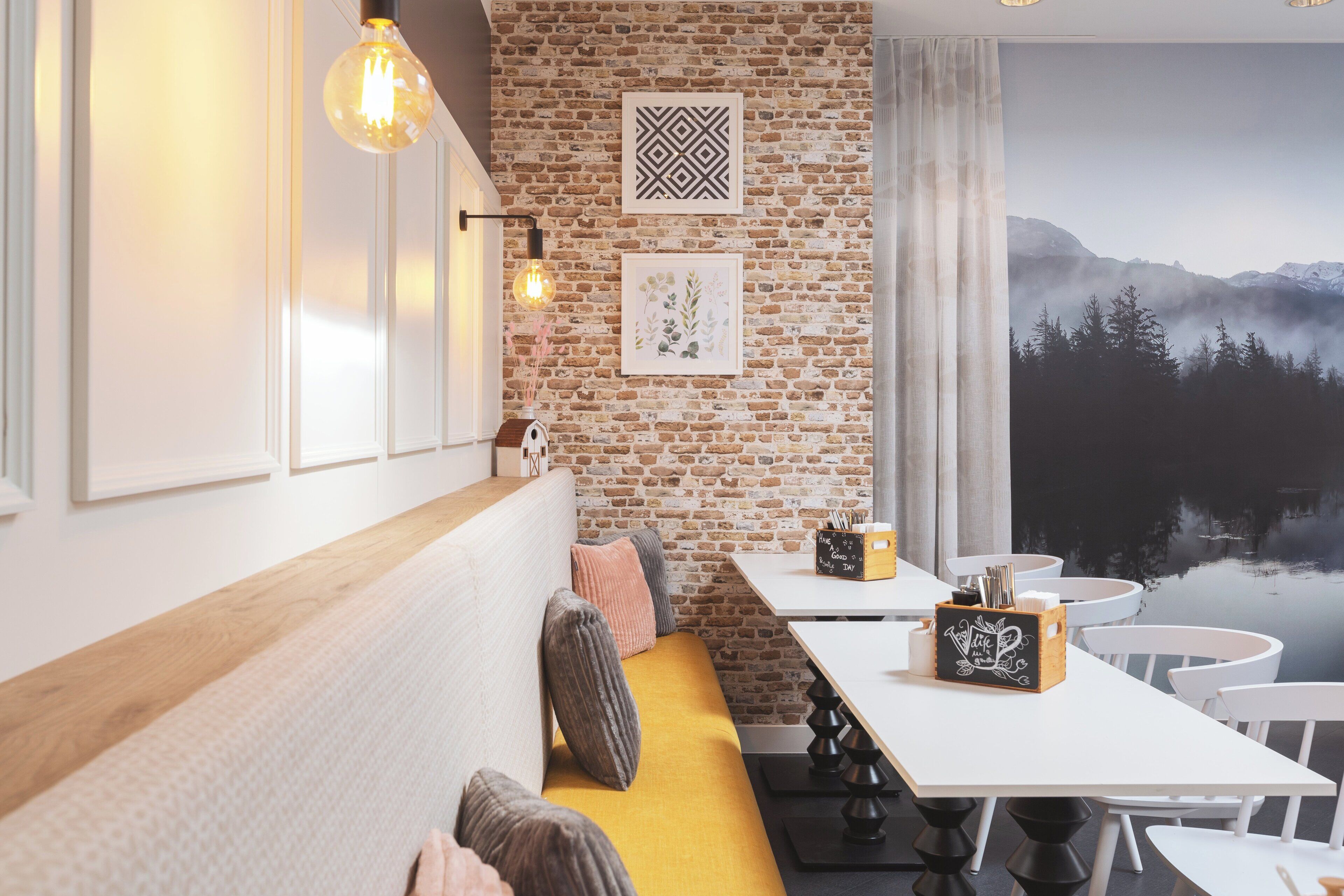 Een gezellige zithoek met een houten bank en kussens, een witte tafel met een schoolbord, en een bakstenen muur met ingelijste kunstwerken.