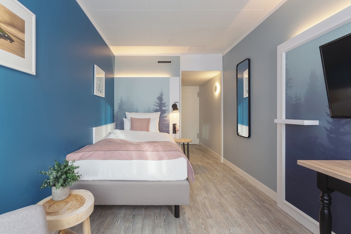 Quarto moderno com uma cama, mesas de cabeceira, uma pequena mesa redonda com uma planta e uma grande janela com vista para árvores.