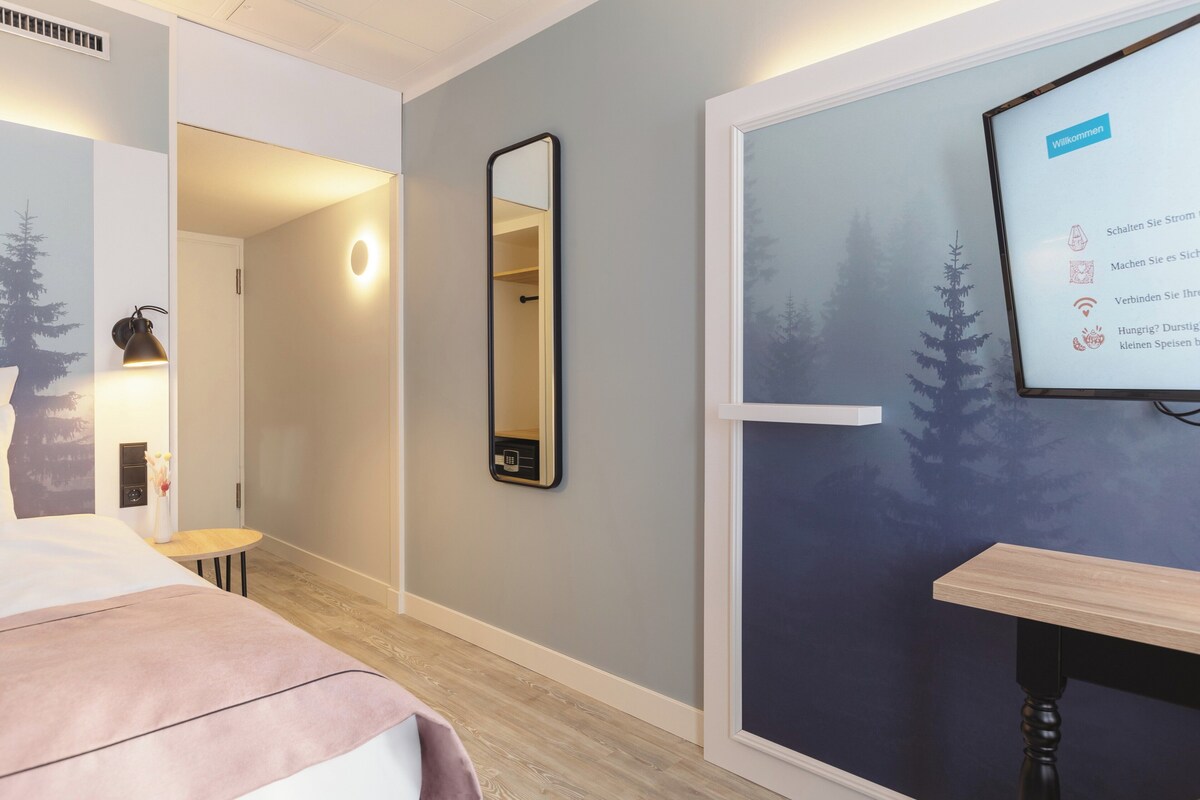 Quarto de hotel com cama, espelho, mesinha e uma tela na parede exibindo mensagens de boas-vindas.