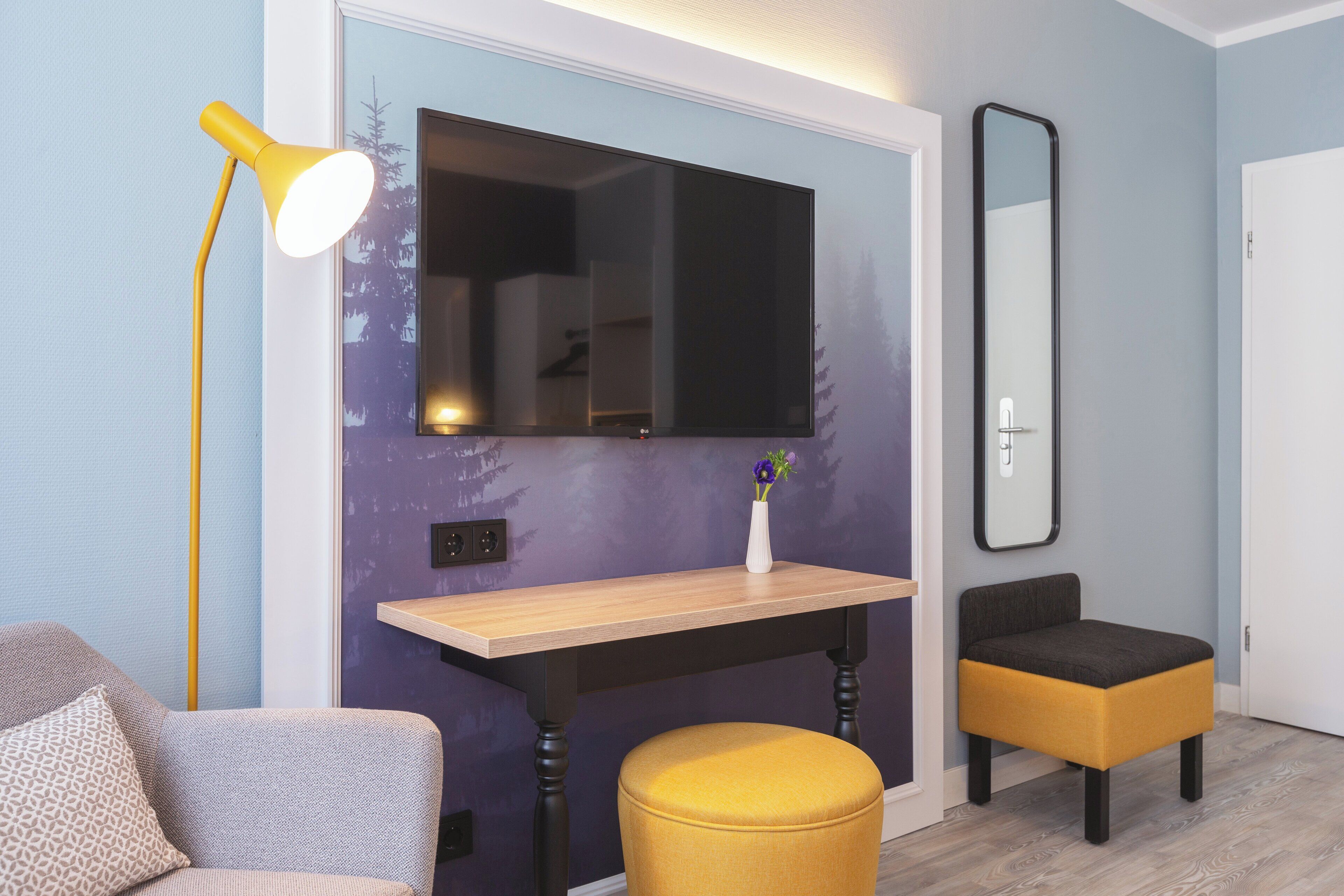 Ein modernes Zimmer mit einem Holztisch, einer gelben Lampe, einem kleinen Tisch mit einer Vase, einem Spiegel und einem grauen Sofa.