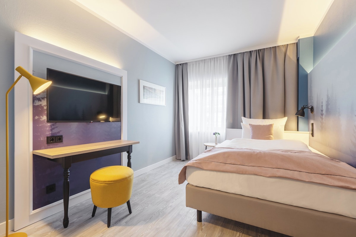 Quarto de hotel moderno com cama, escrivaninha, cadeira e televisão.