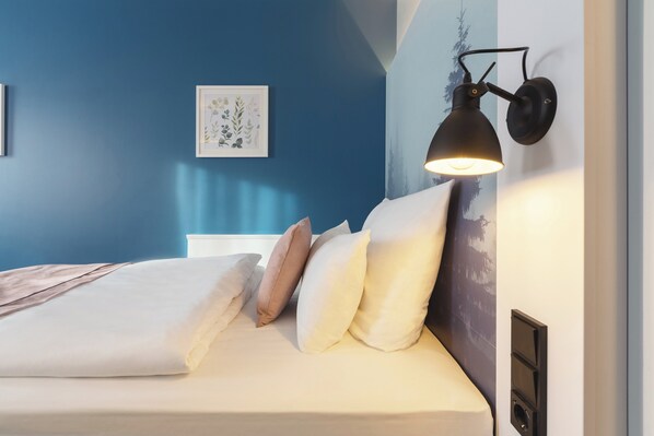A neatly made bed with white and beige bedding, a wall-mounted black lamp, and a framed botanical print on the blue wall. - Garner Hotel Essen - Messe by IHG (Essen)