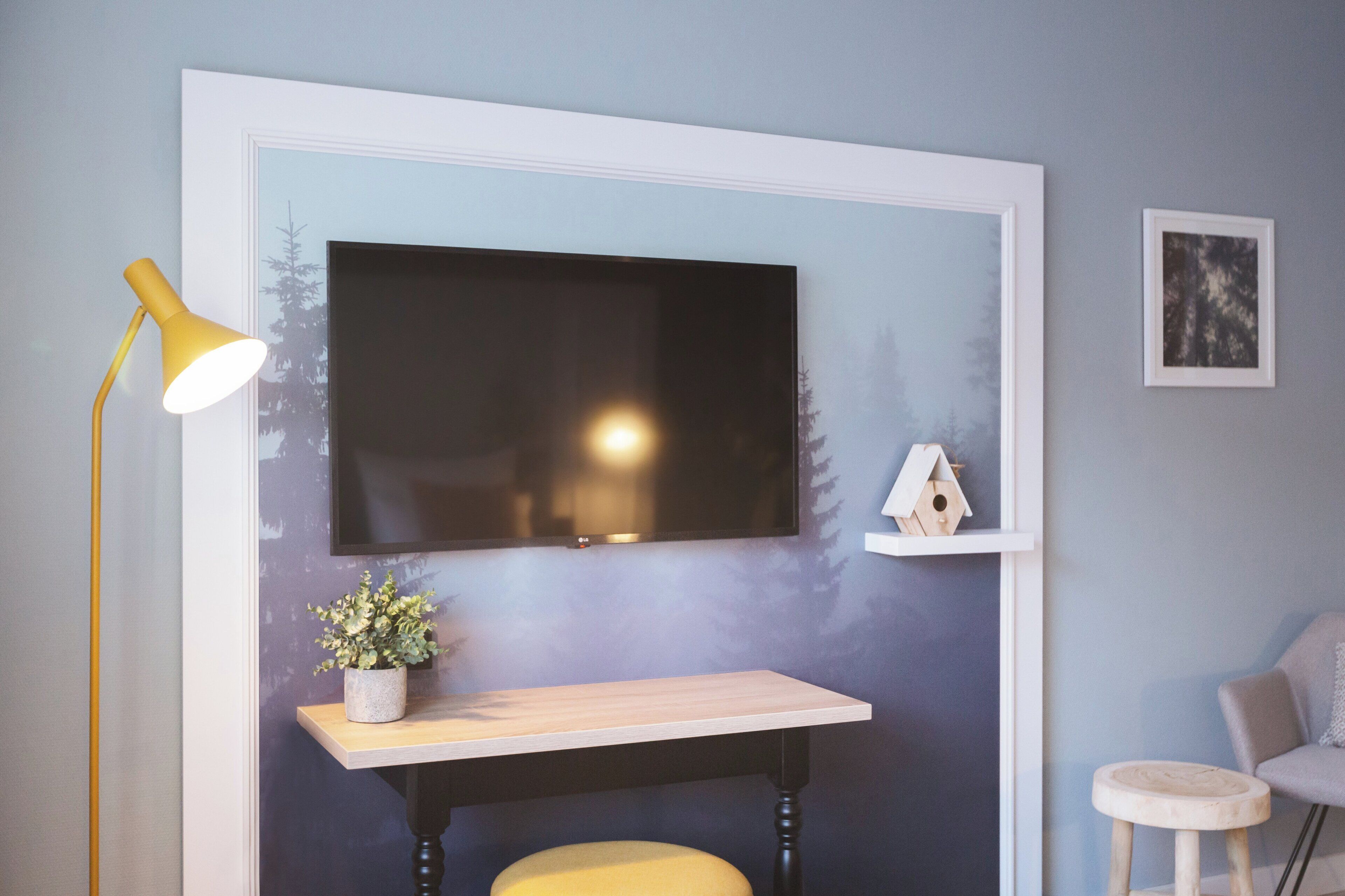 Een kamer met een wandtelevisie, een bureau met een plant, een gele kruk, een lamp en een stoel.