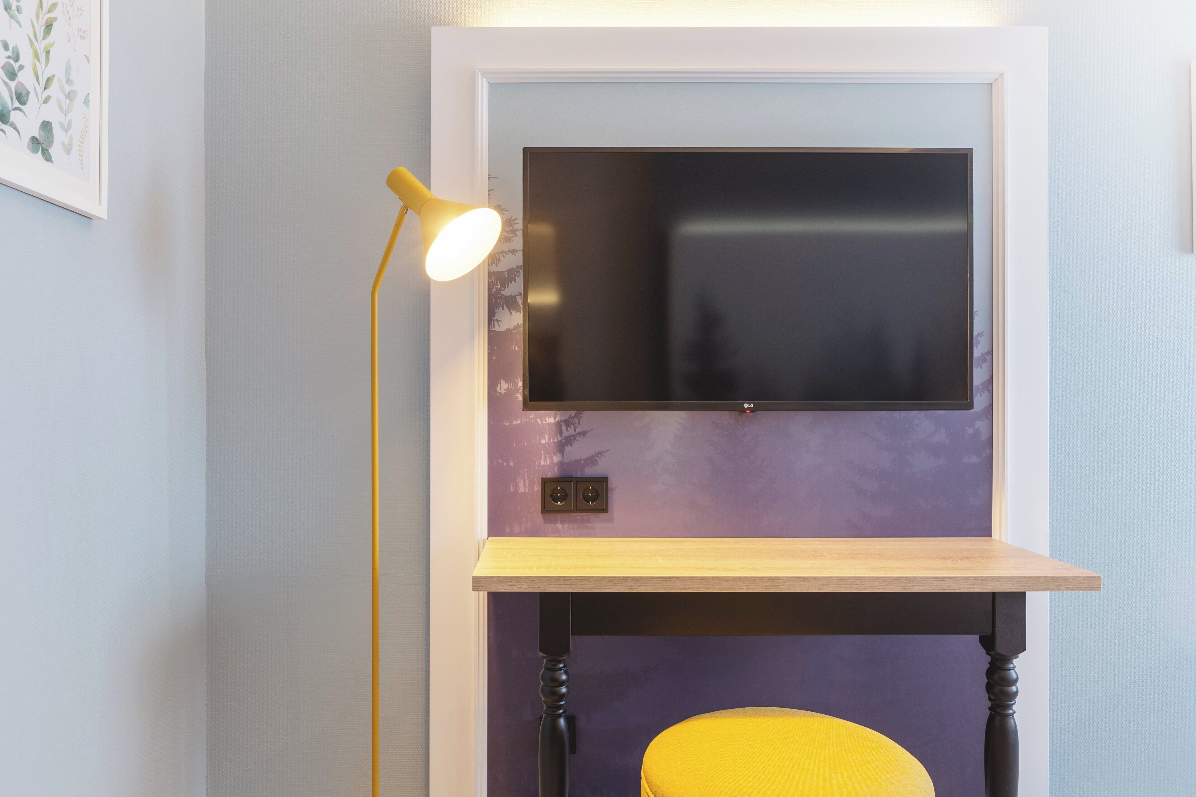 Une pièce avec un mur violet, un téléviseur à écran plat fixé au mur, une étagère en bois, une chaise jaune et une lampe de bureau jaune.