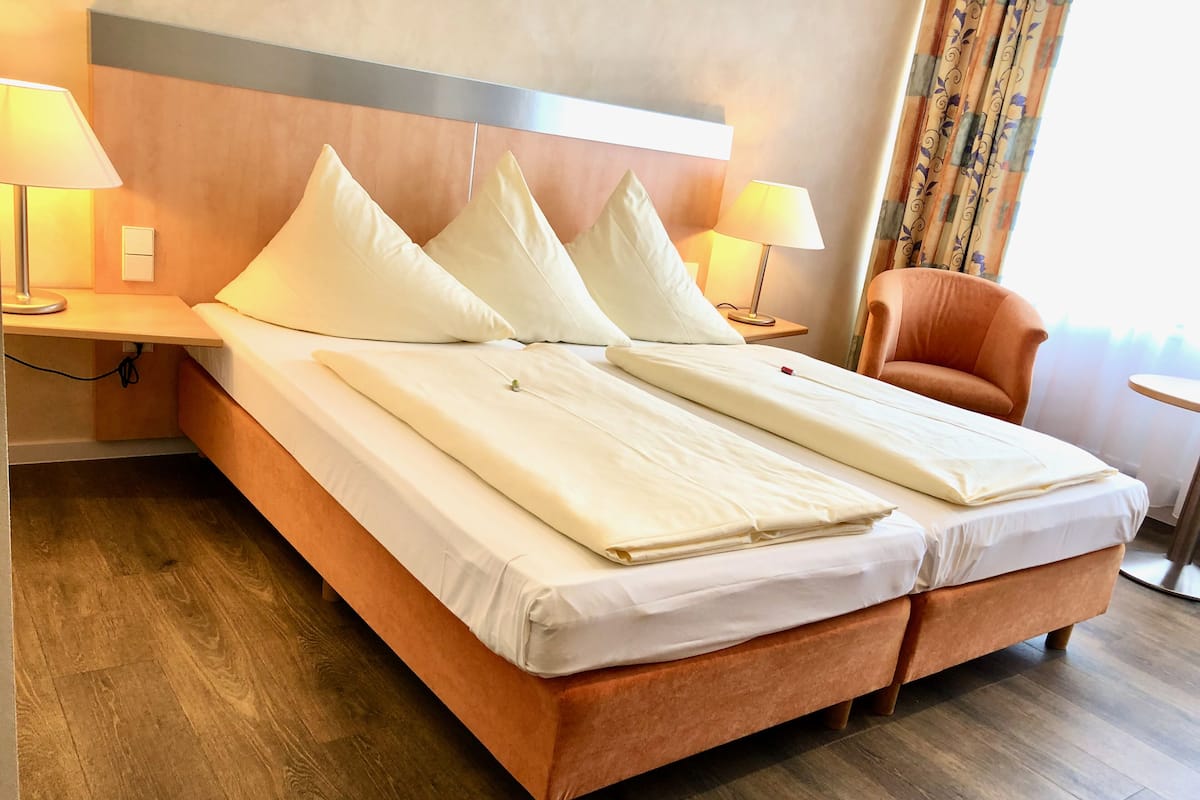 A hotel room with a double bed, two bedside tables with lamps, a wooden headboard, a wooden floor, and a chair.