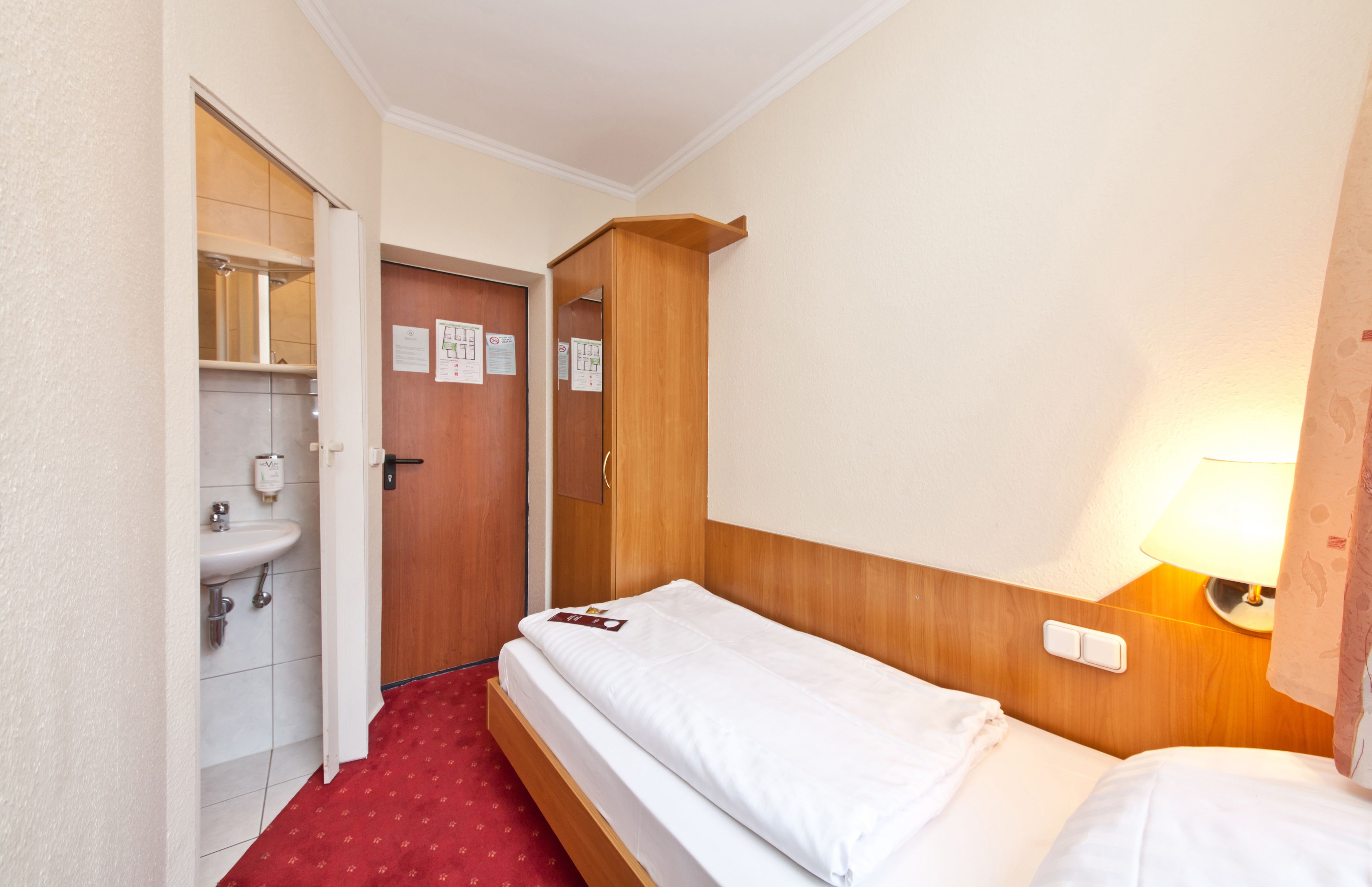 A hotel room with a single bed, a wooden headboard, a bedside lamp, a red carpet, a white sink, and a door leading to a bathroom.