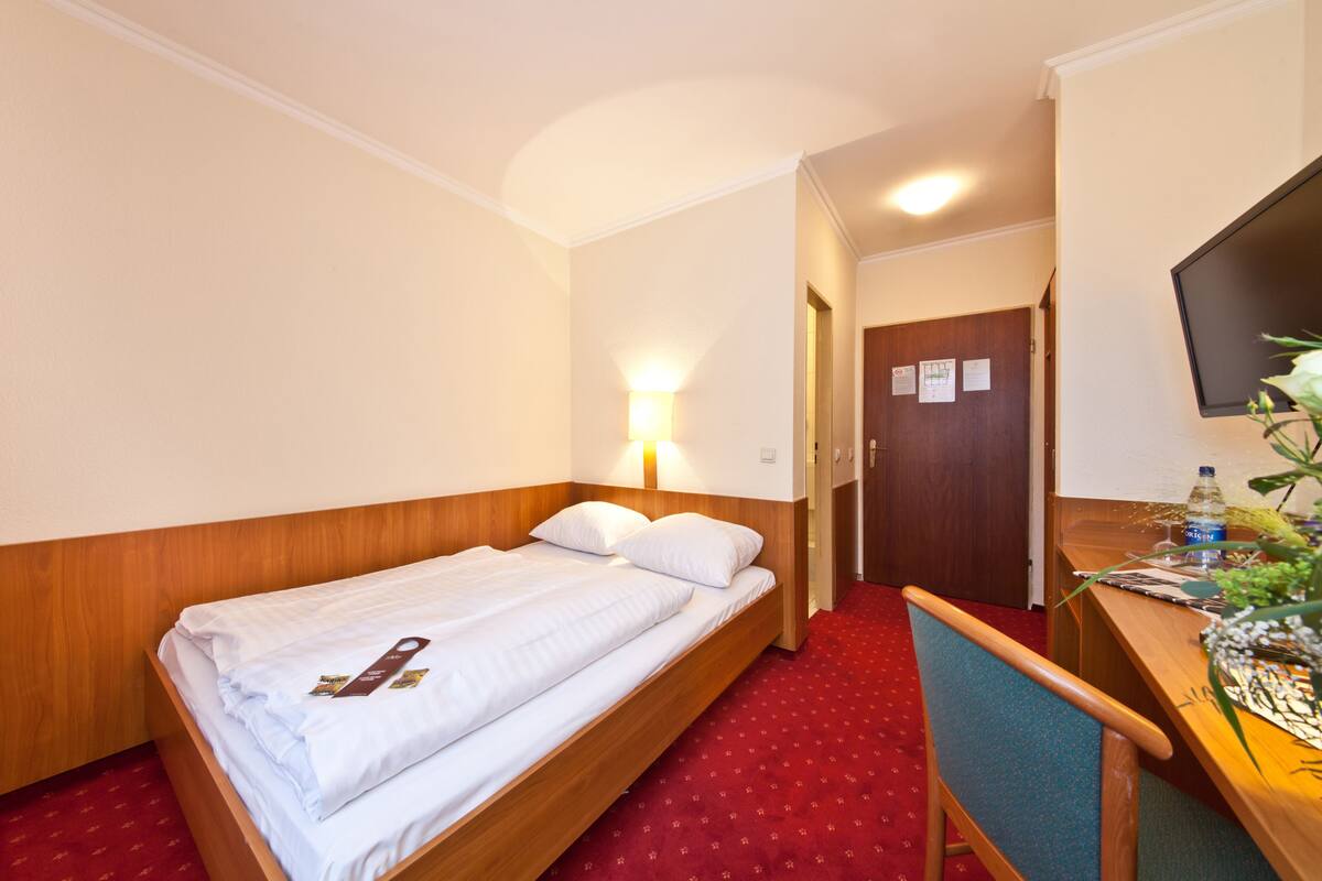 A hotel room with a single bed, a desk with a chair, a lamp, and a TV mounted on the wall.