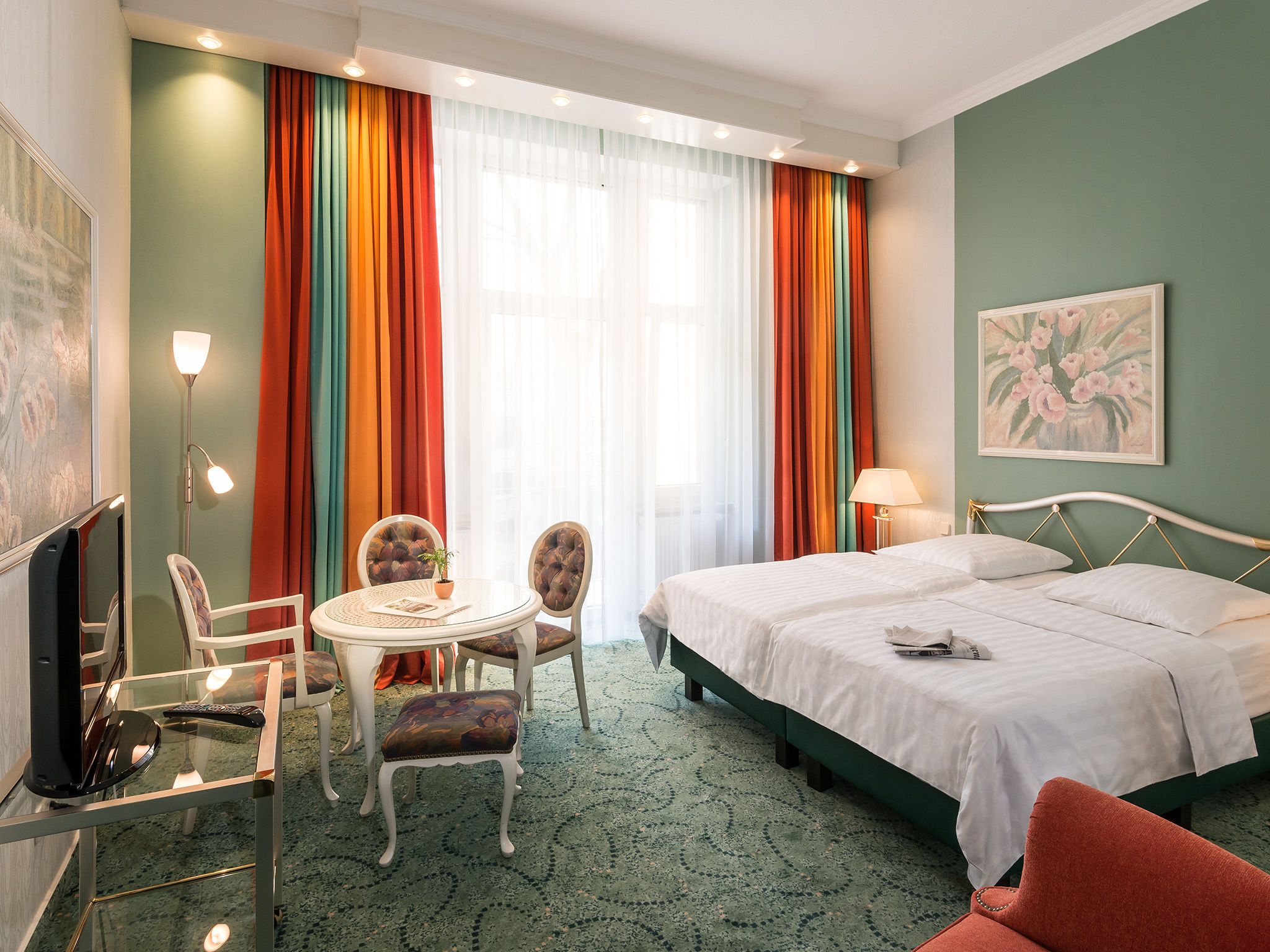 A hotel room with a bed, two chairs, a small table, a TV, and a painting on the wall.