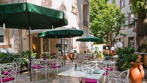 Ein Außenbereich mit einer Terrasse, weißen Korbmöbeln, grünen Sonnenschirmen und Terrakottatöpfen.