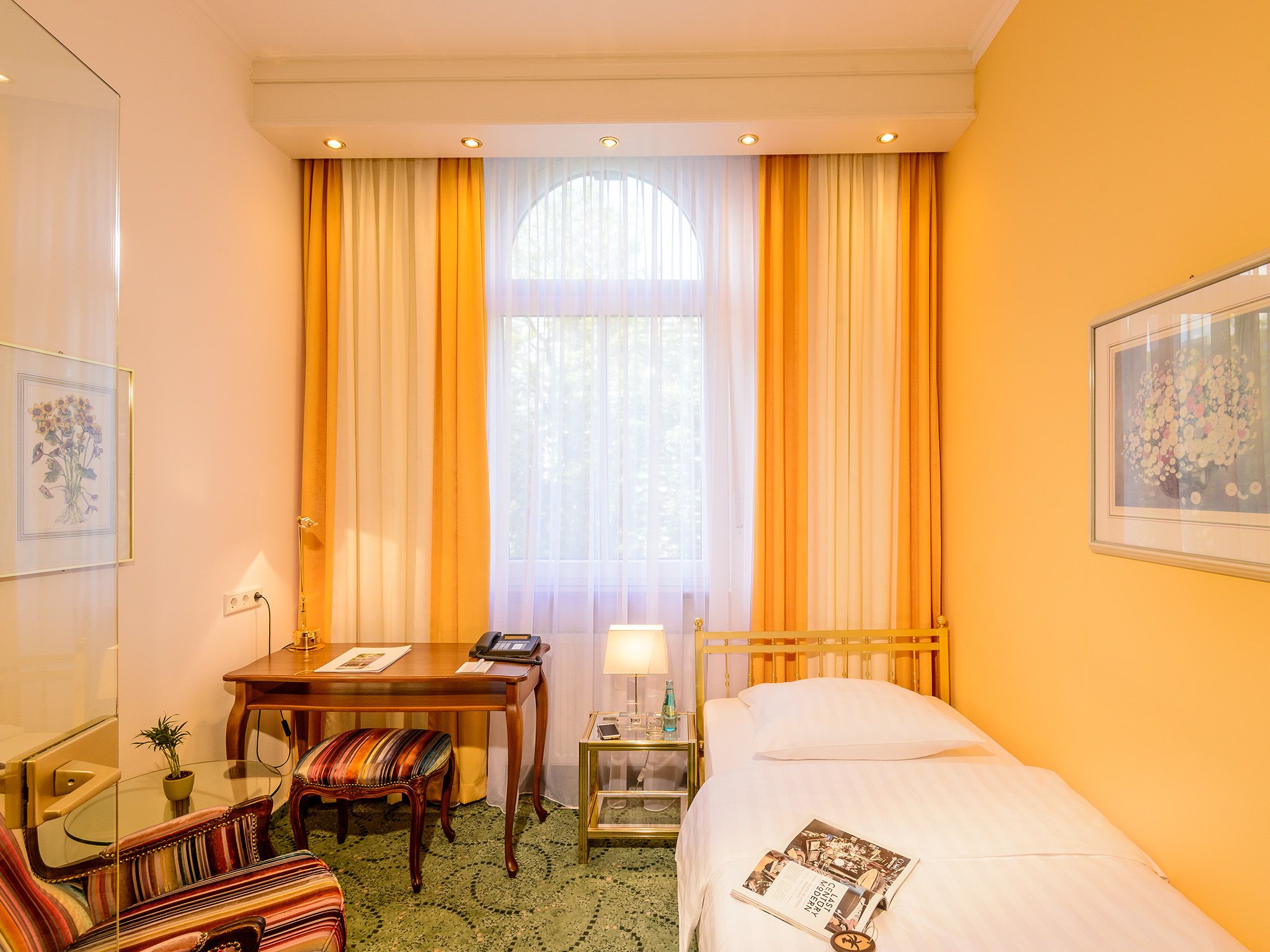 A hotel room with a bed, a desk, a chair, a small table, and a window with curtains.