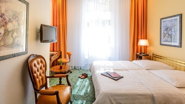 Une chambre d’hôtel avec un lit, un bureau, une chaise, une télévision et une fenêtre avec des rideaux.