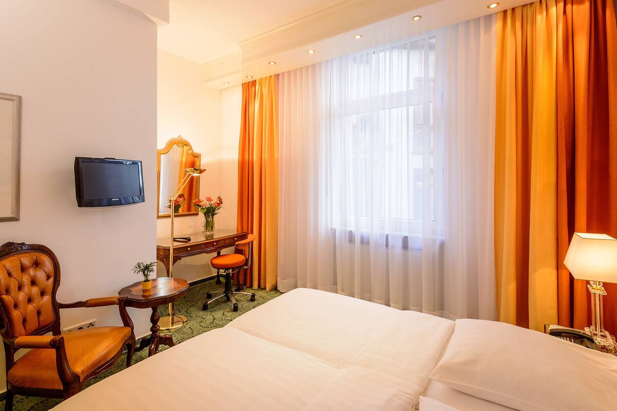 Ein Hotelzimmer mit Bett, Schreibtisch mit Stuhl, Fernseher, Spiegel und einem Fenster mit Vorhängen.