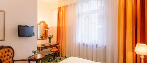 A hotel room with a bed, a desk with a chair, a television, a mirror, and a window with curtains.