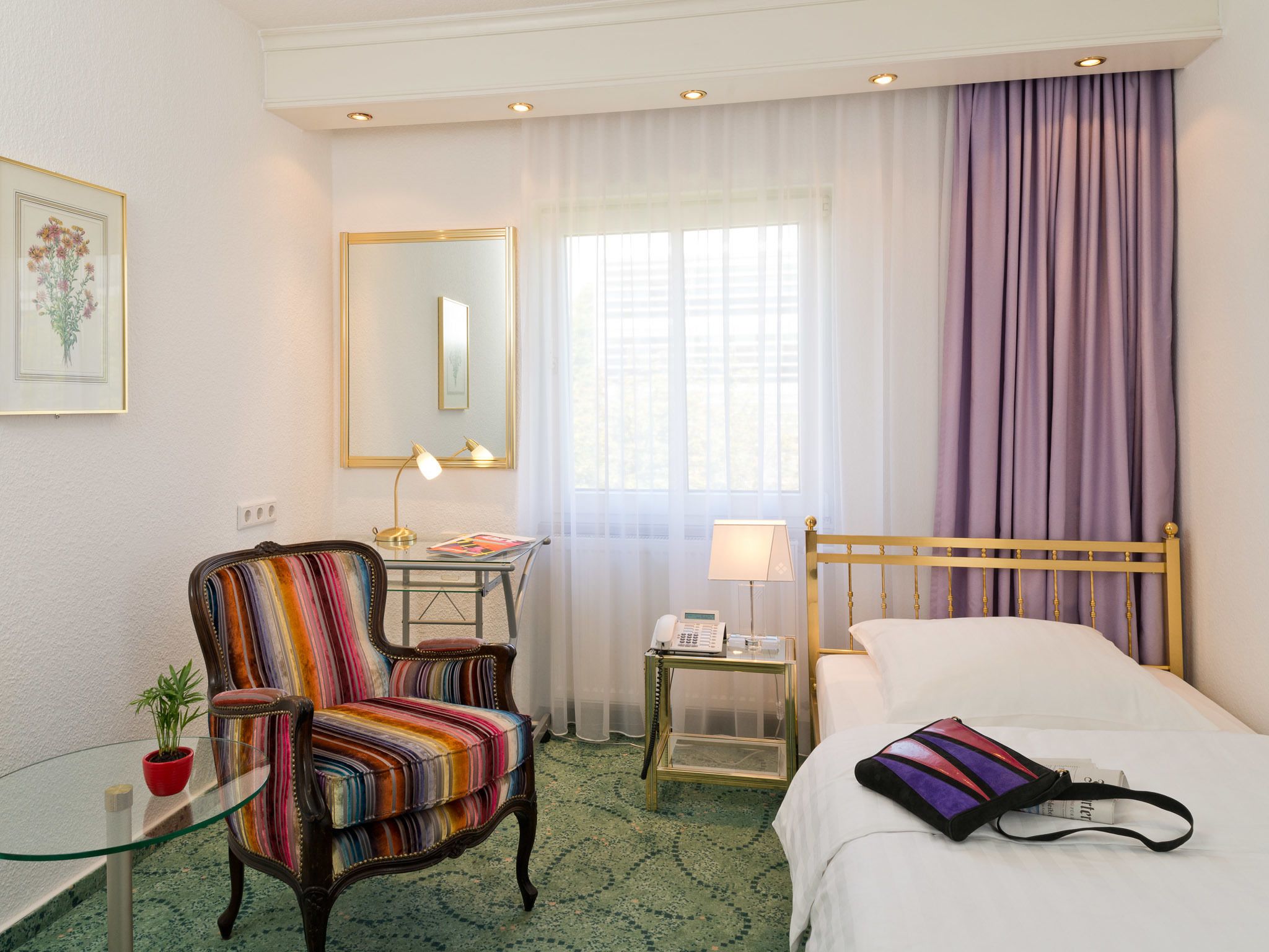 A hotel room with a bed, a striped armchair, a small table with a lamp, a bedside table with a phone, a window with curtains, and a mirror on the wall.