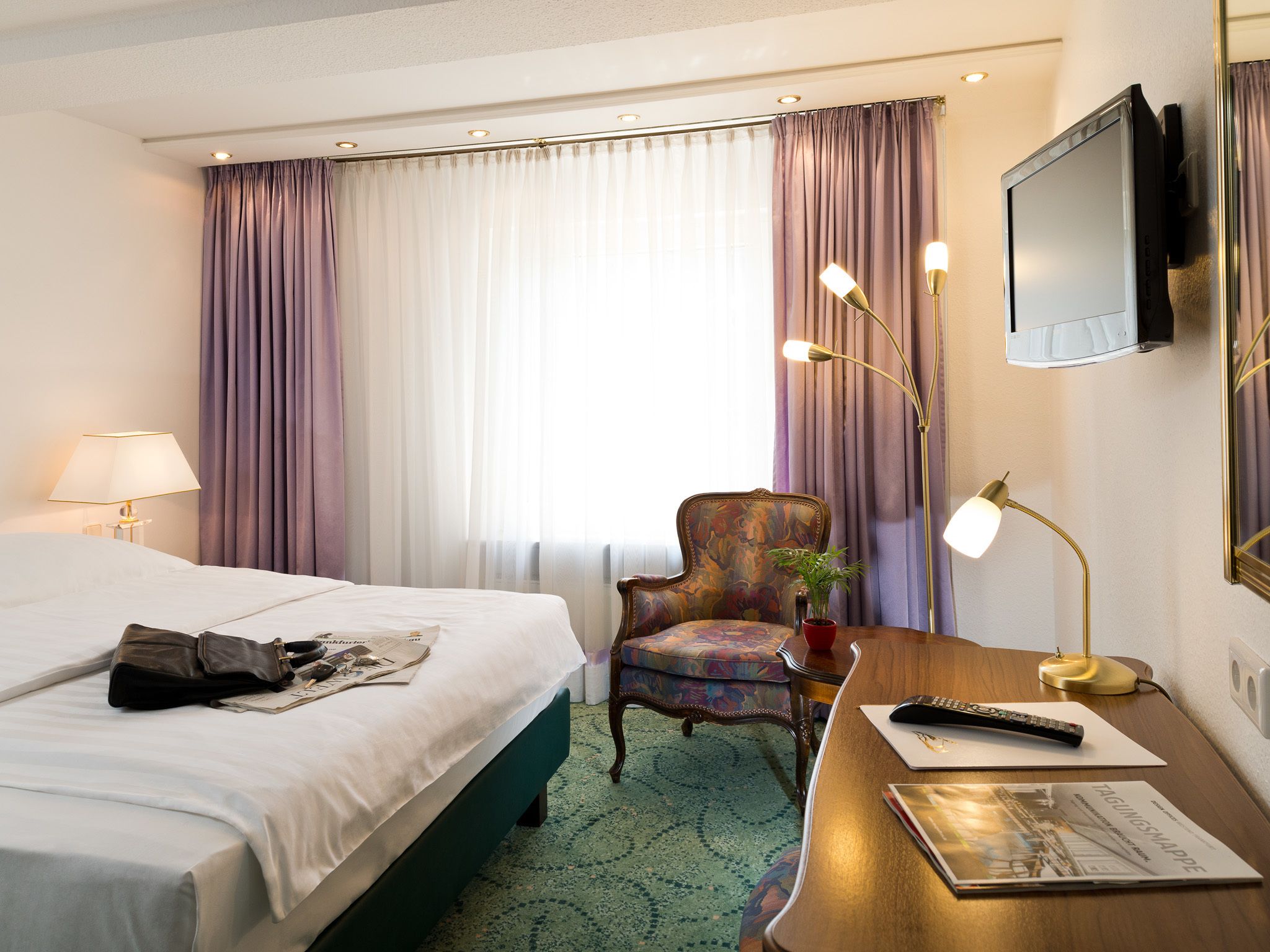 A hotel room with a bed, a desk with a lamp, a chair, and a television mounted on the wall.