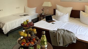 Hotellihuone, jossa on kaksi erillistä sänkyä, ruokapöytä, jonka päällä on hedelmiä ja pullo, kannettava tietokone suuremmalla sängyllä ja taulu seinällä.