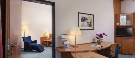 A hotel room with a wooden desk, a chair, a TV, a lamp, a framed picture, and a small table with a vase of flowers.