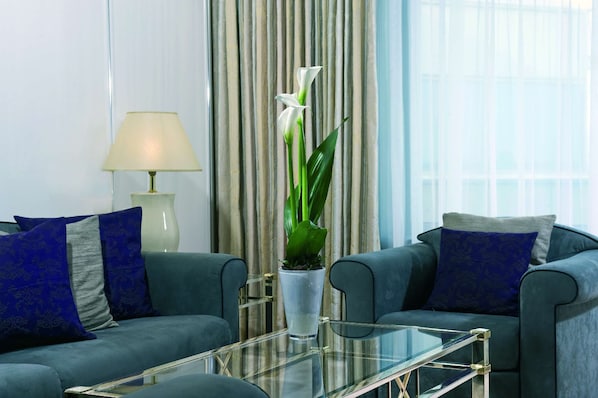 Una sala de estar moderna con una mesa de centro de vidrio, dos sofás grises con cojines azules, un jarrón blanco alto con una planta y una lámpara.