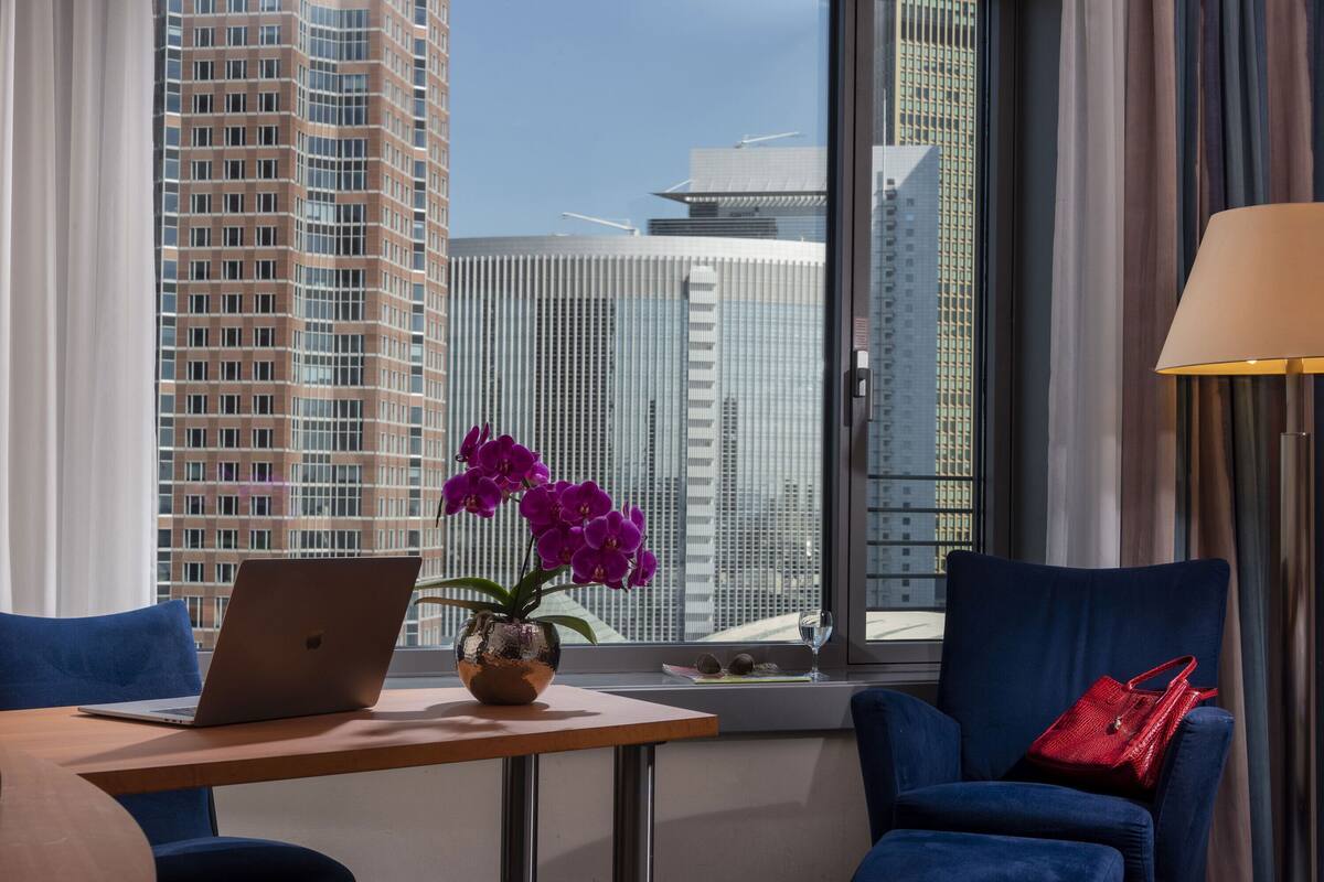 Een moderne hotelkamer met een laptop op een tafel, een vaas met paarse bloemen, een rode handtas en uitzicht op hoge gebouwen.