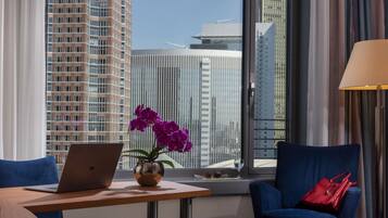 A modern hotel room with a laptop on a table, a vase with purple flowers, a red handbag, and a view of tall buildings.