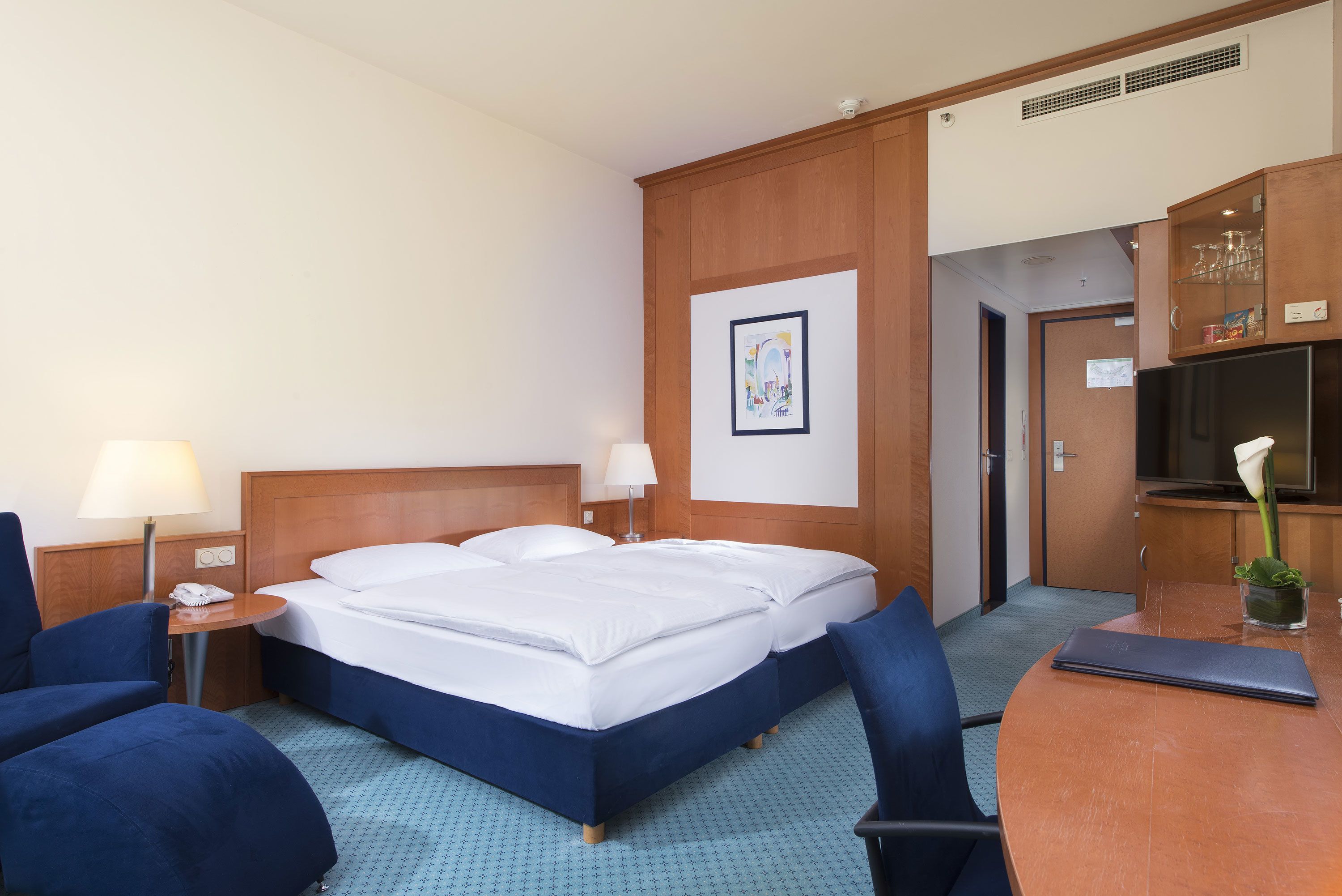 A hotel room with a double bed, two bedside tables with lamps, a desk with a laptop, a chair, a small table with a vase, and a door leading to a bathroom.