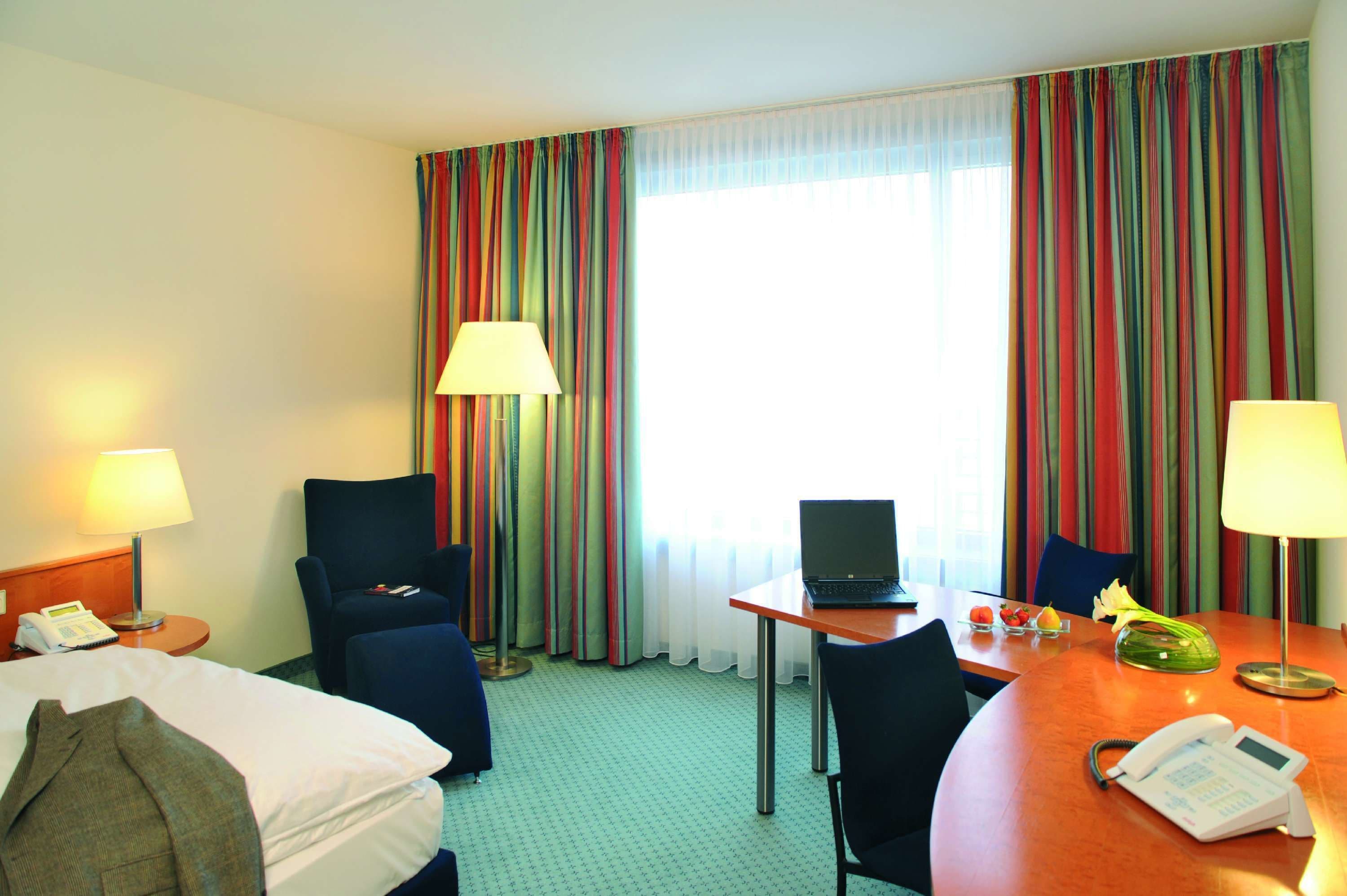 A hotel room with a bed, a desk with a laptop, a chair, two lamps, a telephone, and a window with striped curtains.