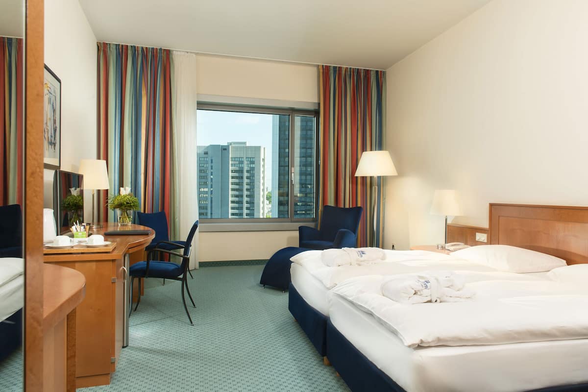 A hotel room with a large bed, a desk with a chair, a small table with a vase, and a window with curtains.