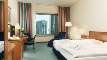 A hotel room with a large bed, a desk with a chair, a small table with a vase, and a window with curtains.