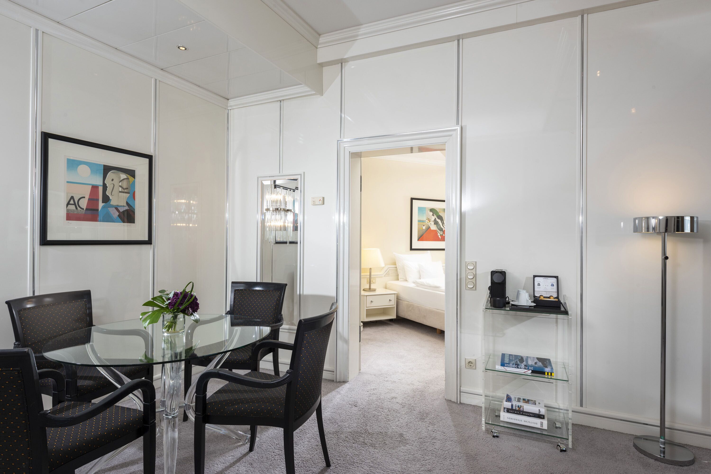 Une chambre d’hôtel moderne avec une table à manger en verre, des chaises noires, un petit vase avec des fleurs, un canapé blanc, une table de chevet avec une lampe et une bibliothèque contenant des livres et un certificat encadré.