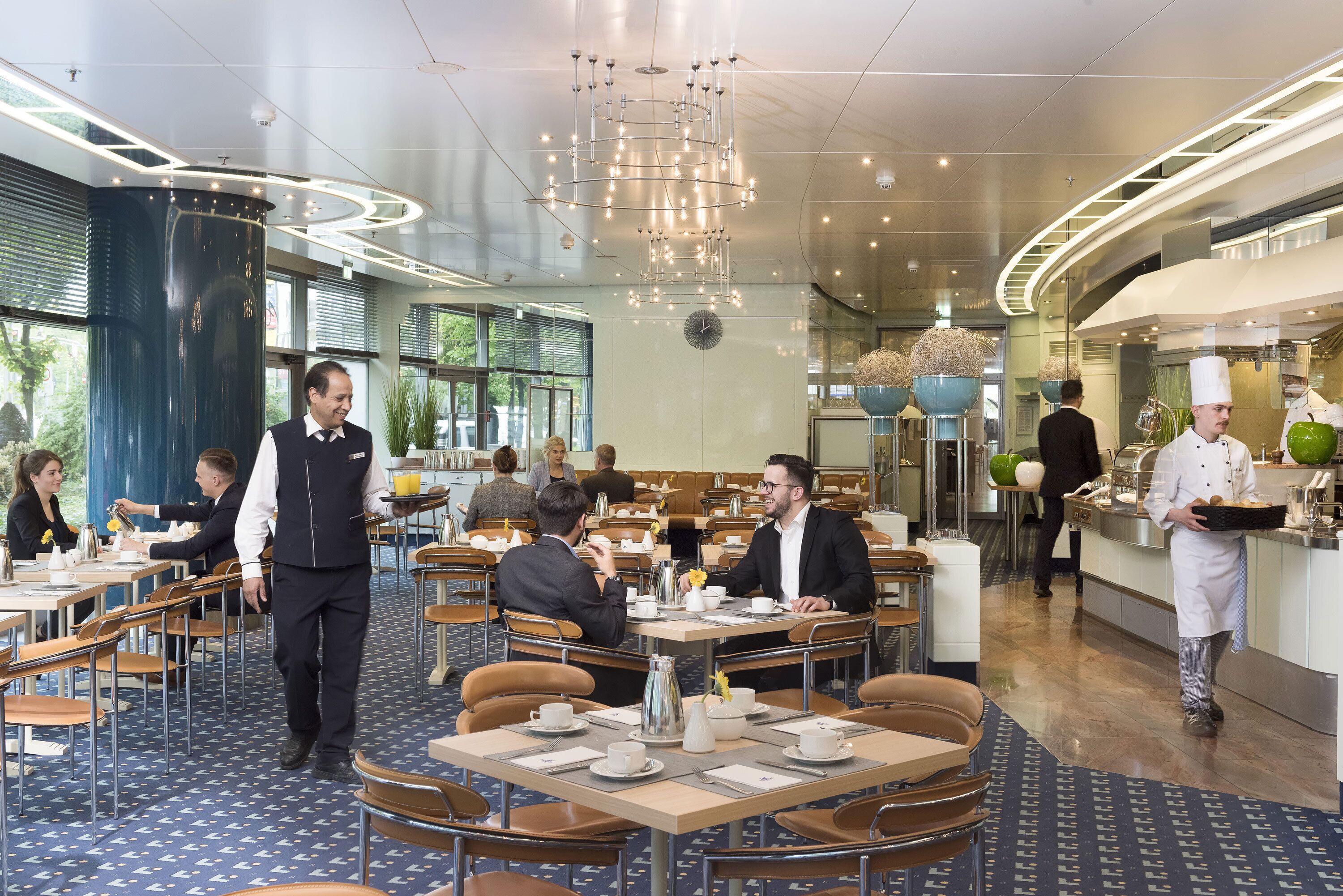 A modern dining area with round tables, a central serving station, and a visible kitchen area.