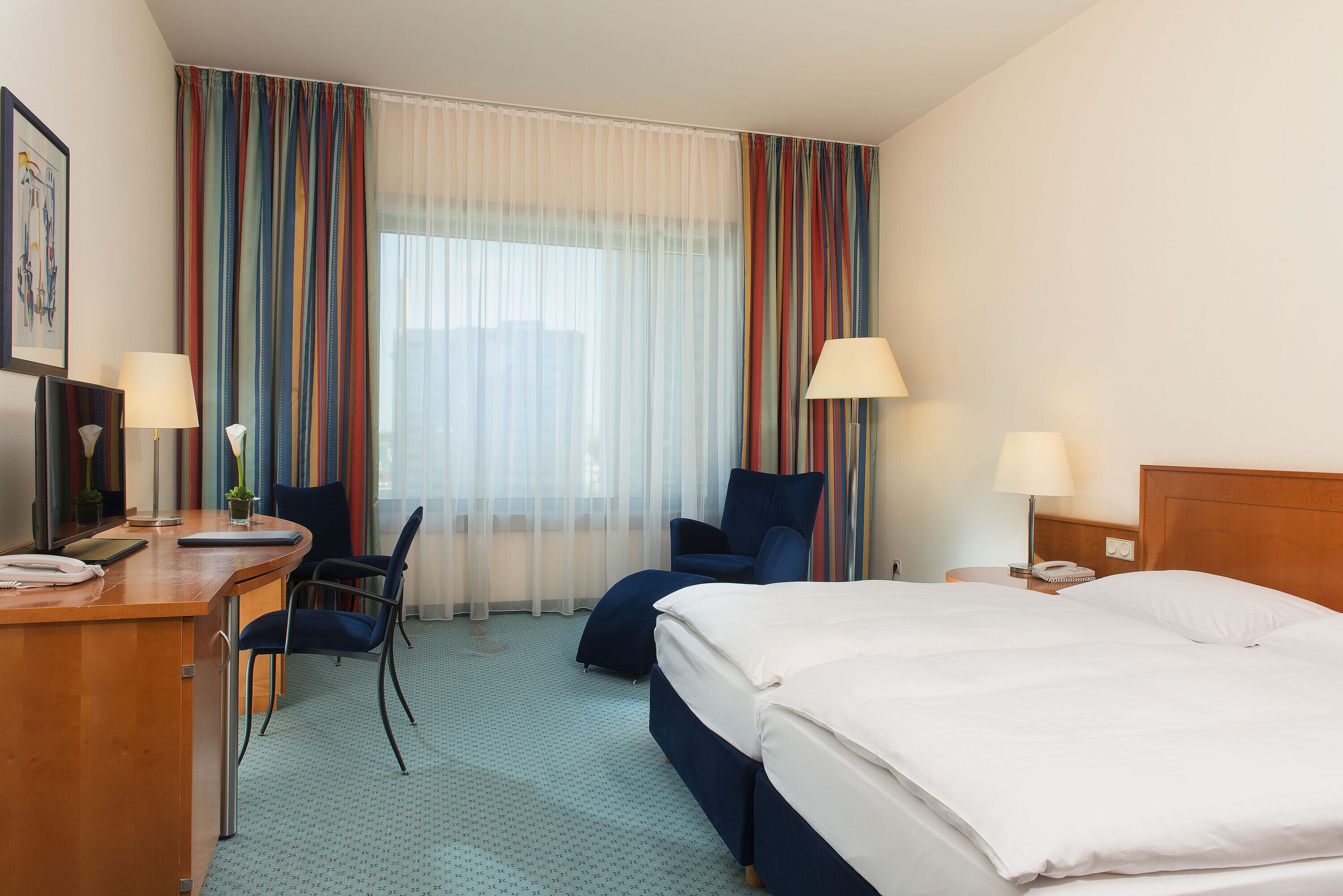 A hotel room with a bed, desk, chair, and a view of the city through large windows.
