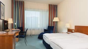 A hotel room with a bed, desk, chair, and a view of the city through large windows.