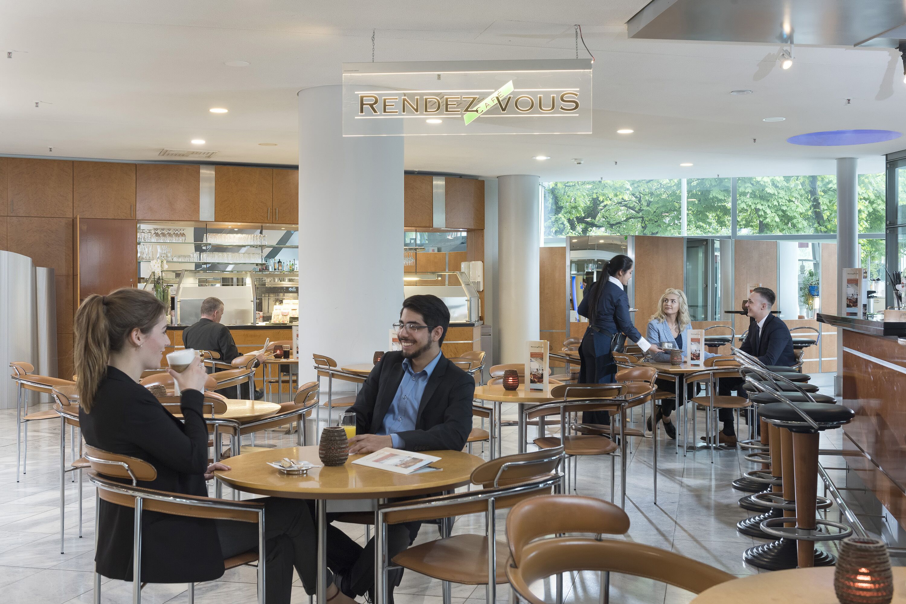 A modern café with wooden tables and chairs, a visible menu board, and a sign reading "RENDEZ VOUX".