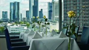 Mesa de comedor dispuesta con manteles blancos y jarrones de flores, con vista al horizonte de una ciudad al fondo.