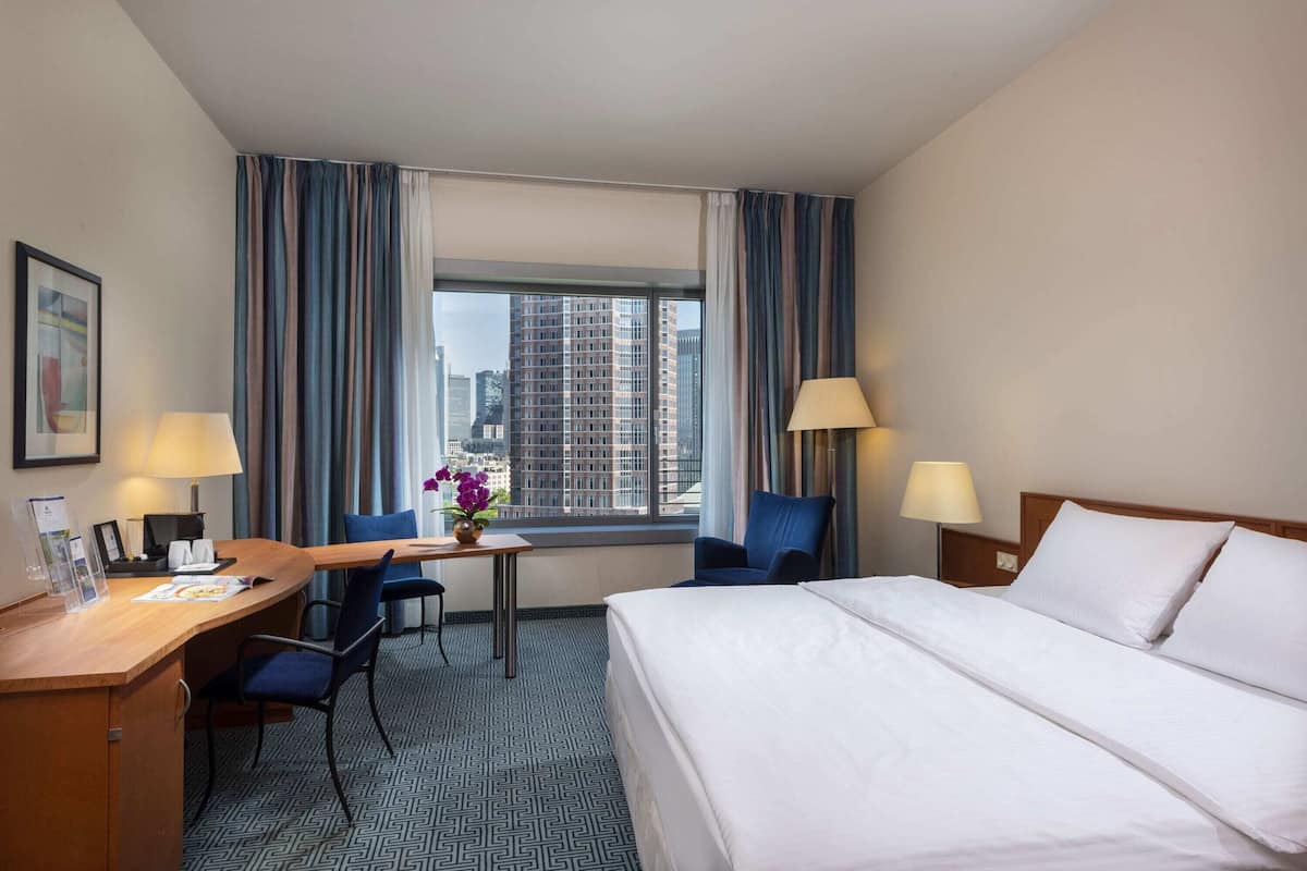 A hotel room with a bed, desk, chairs, lamps, and a window with curtains.