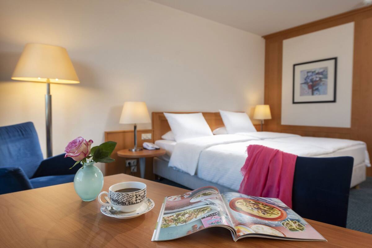 Een hotelkamer met een bed, nachtlampjes, een salontafel met een tijdschrift, een vaas met een bloem en een blauwe fauteuil.