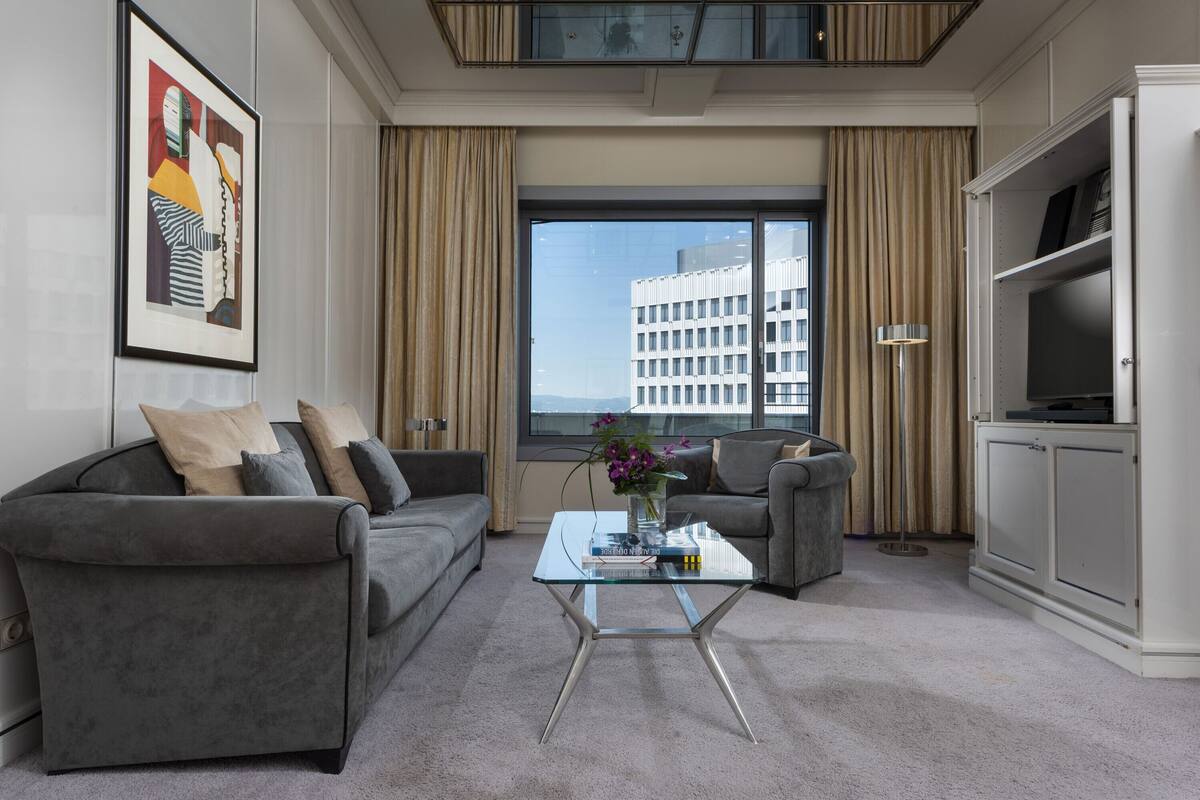 Een moderne woonkamer met een grijze bank, een glazen salontafel en uitzicht op een gebouw door grote ramen.