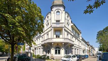 Un bâtiment historique avec une entrée située à l’angle, une fontaine devant et des voitures garées le long de la rue.