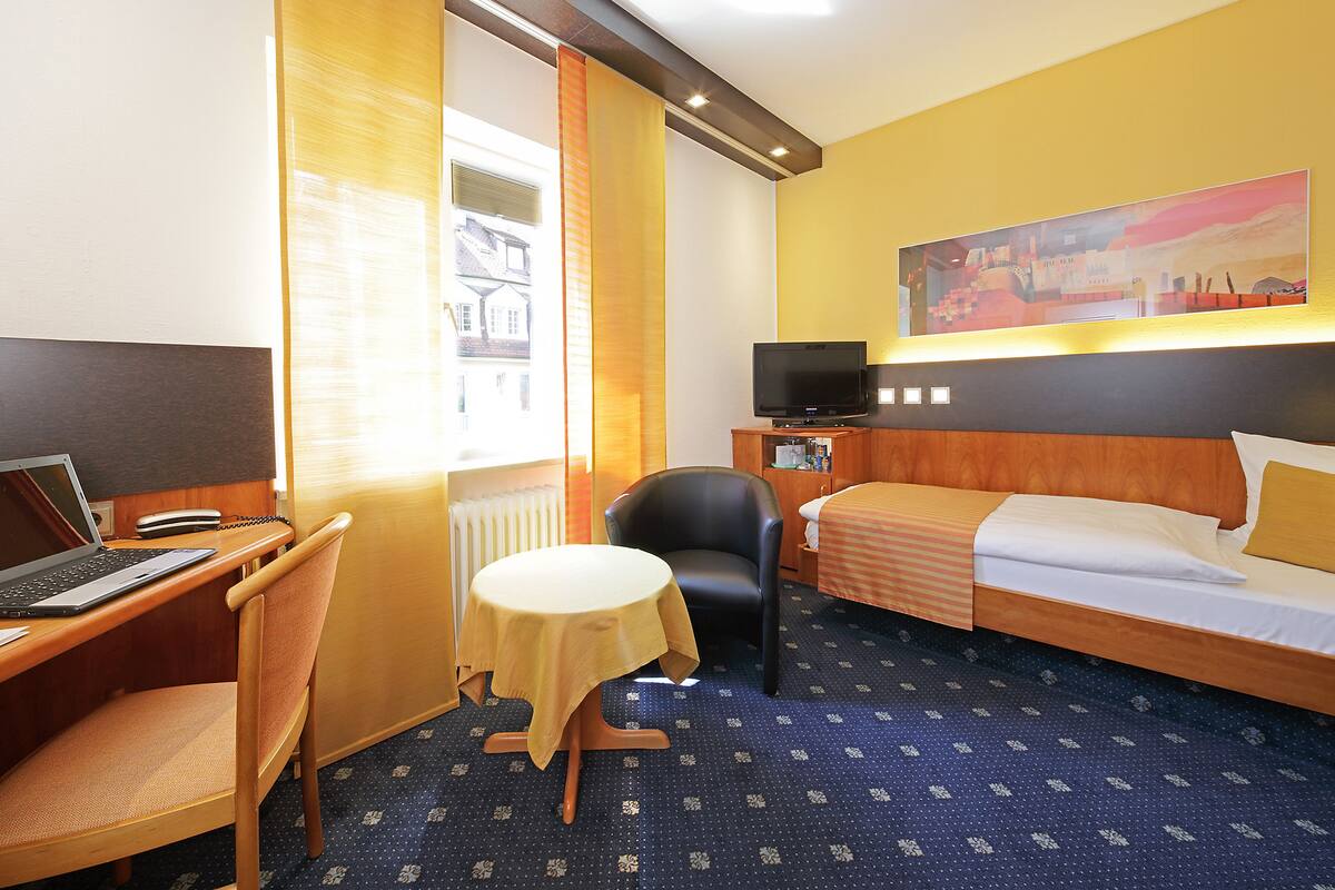 A hotel room with a bed, a desk with a laptop, a chair, a small table, and a TV.