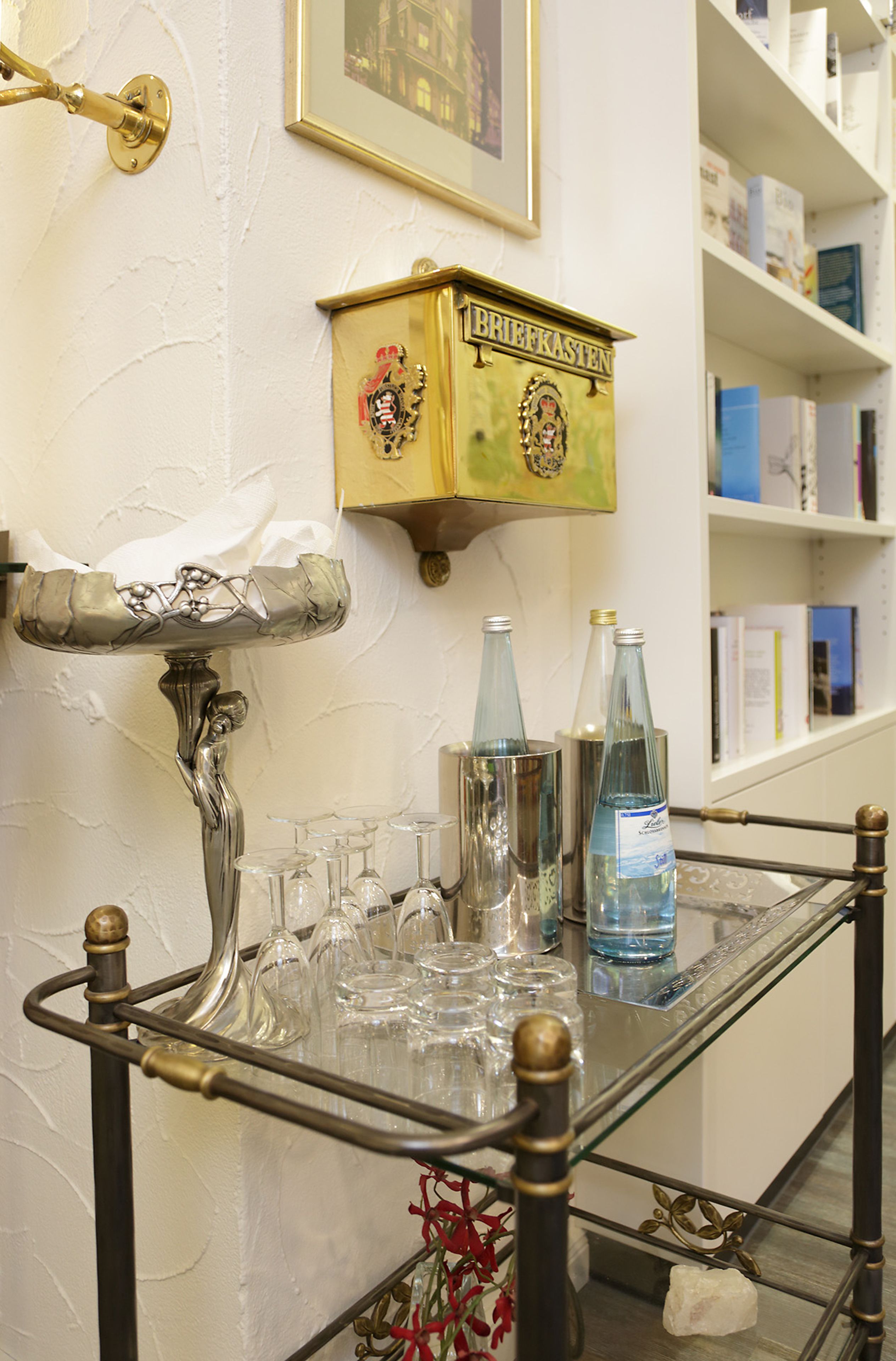 Een bar met een barwagen met glazen blad, een messing lamp en een plank met boeken en flessen.