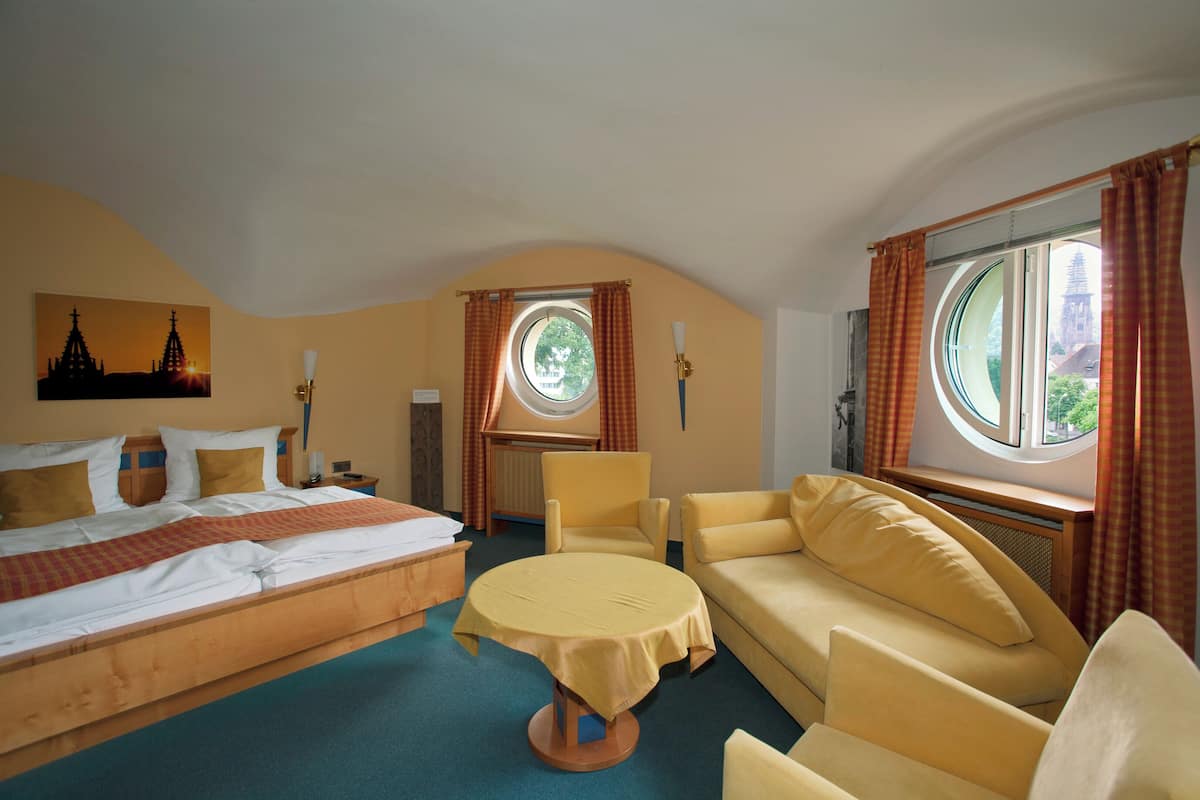 A hotel room with a bed, a round table, a sofa, and two windows with curtains.