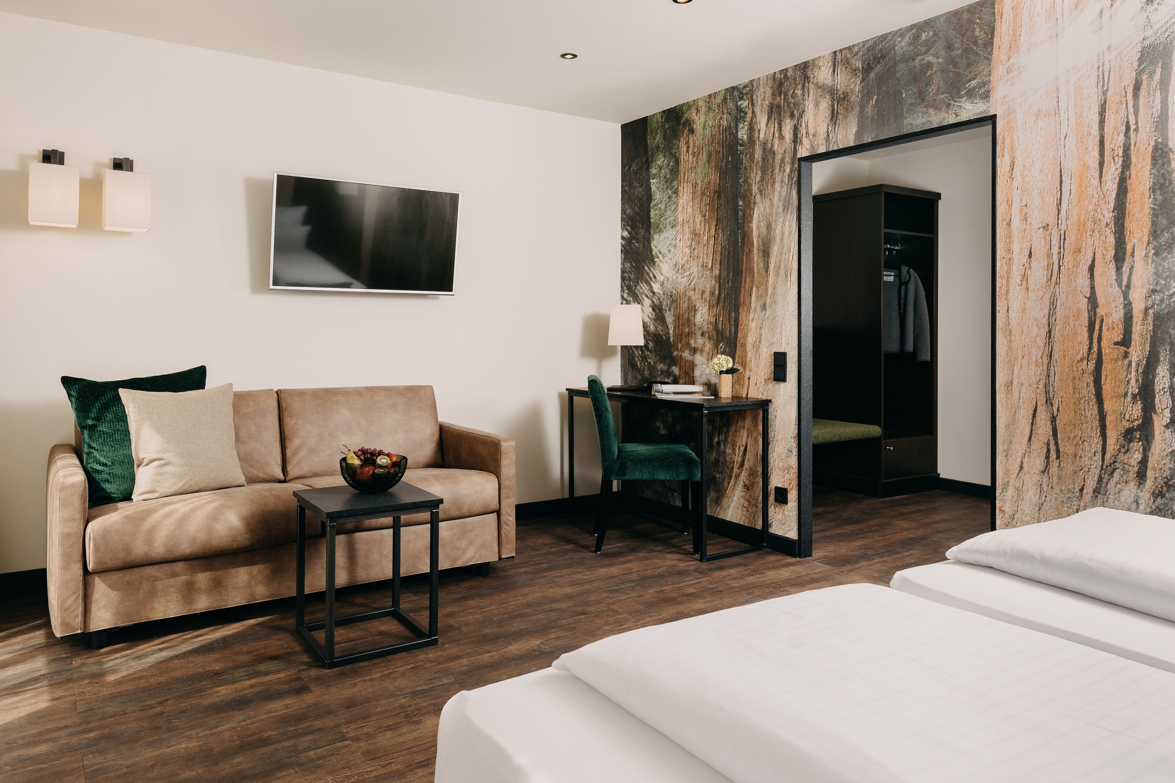 A modern hotel room with a sofa, a small table with a bowl of fruit, a desk with a lamp, and a large mirror.