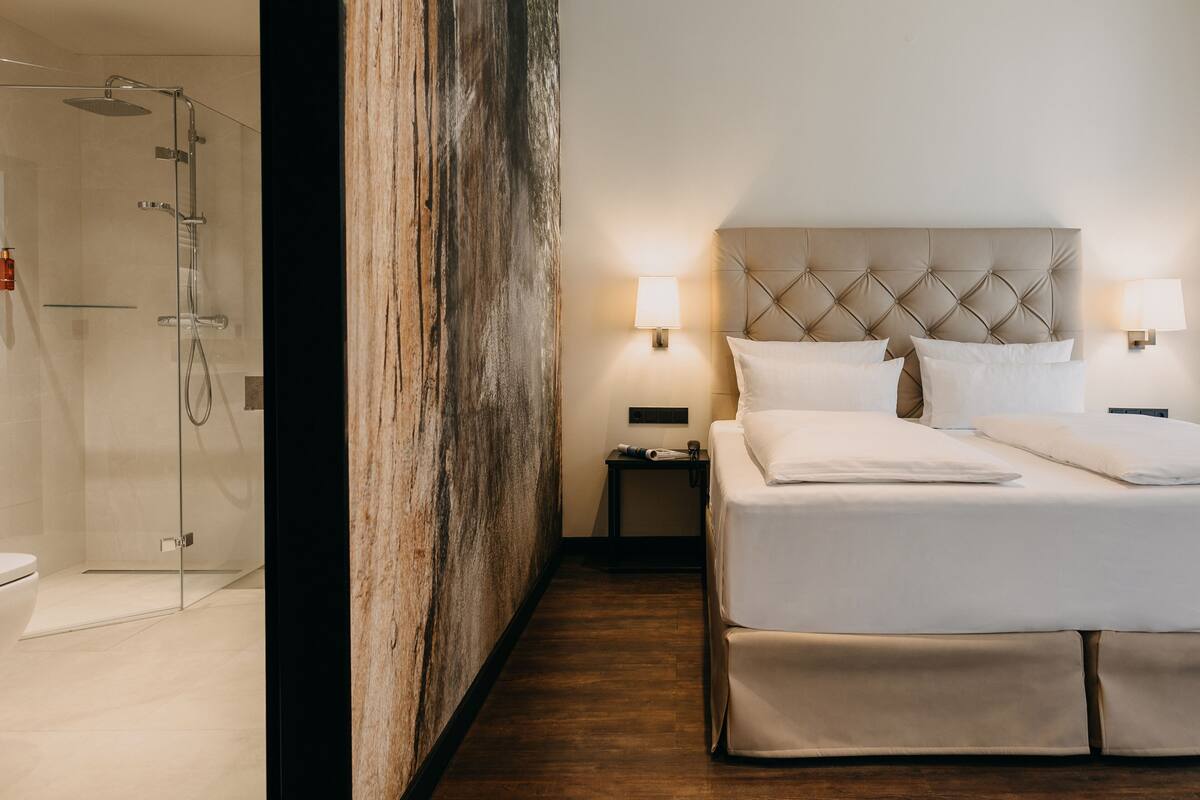 Una camera d'albergo con un letto, lampade da comodino, un comodino e uno spazio doccia.