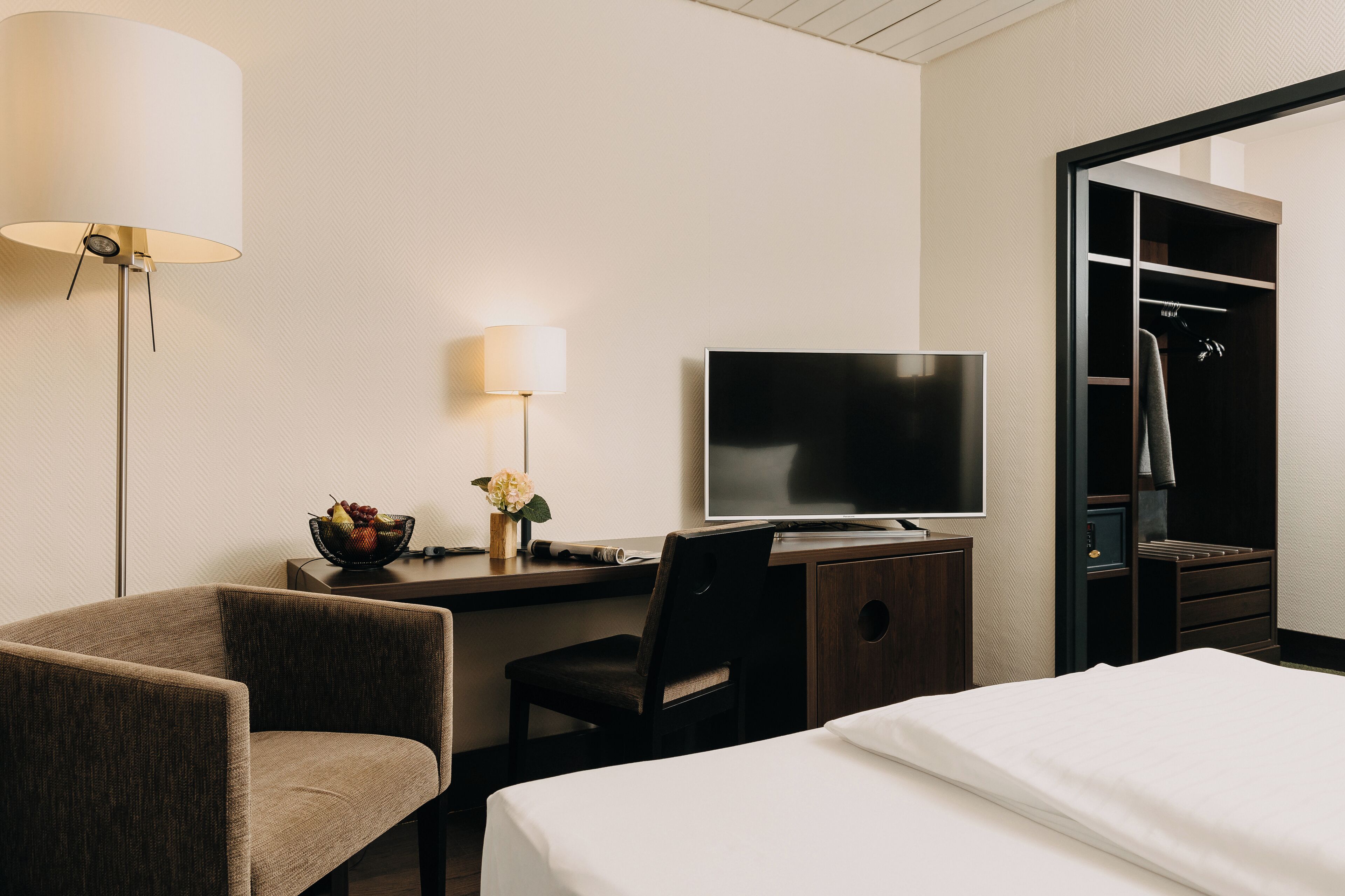 A hotel room with a bed, a desk with a TV, a chair, a lamp, and a wardrobe.