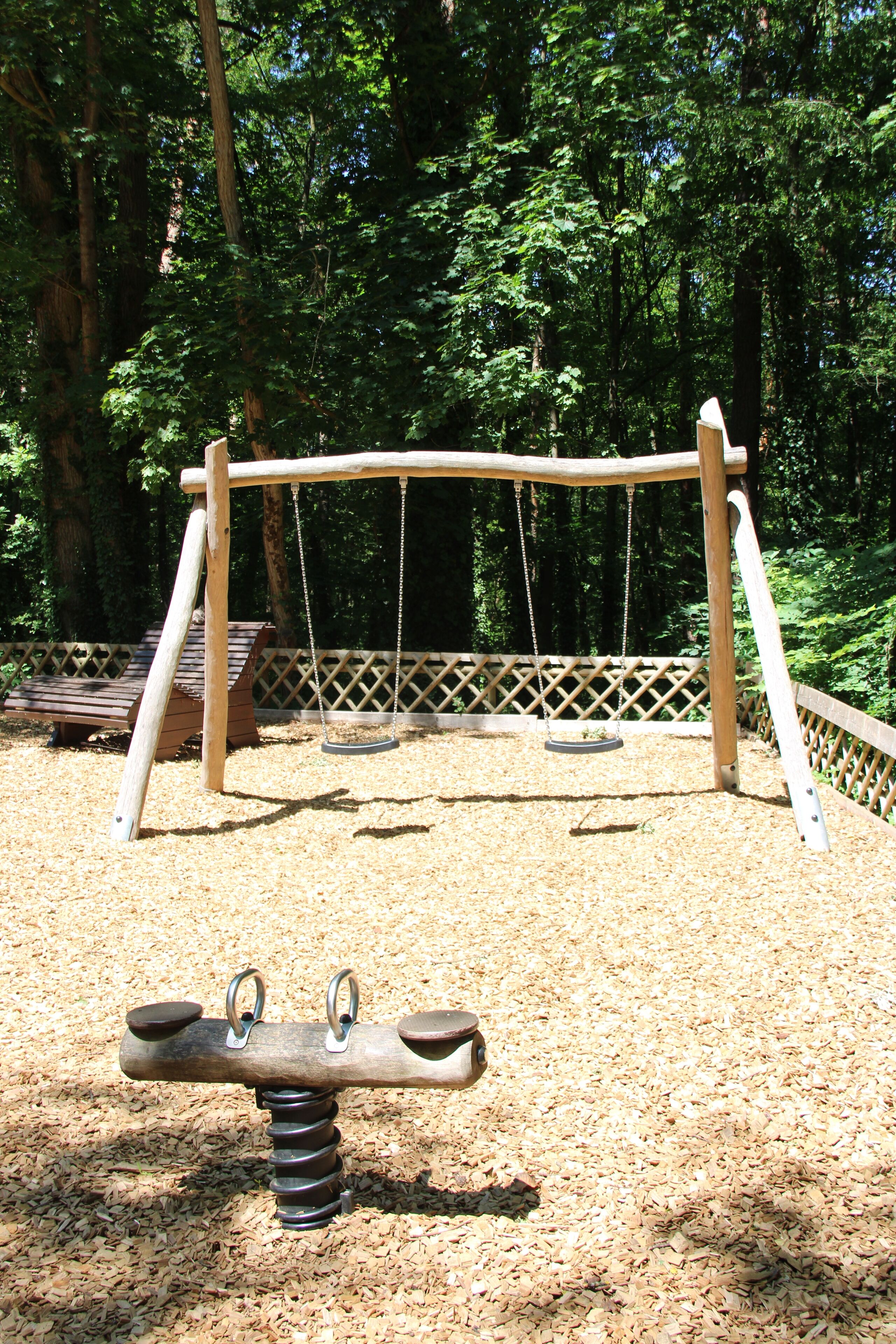 A playground with a seesaw and swings, surrounded by a wooden fence and trees.