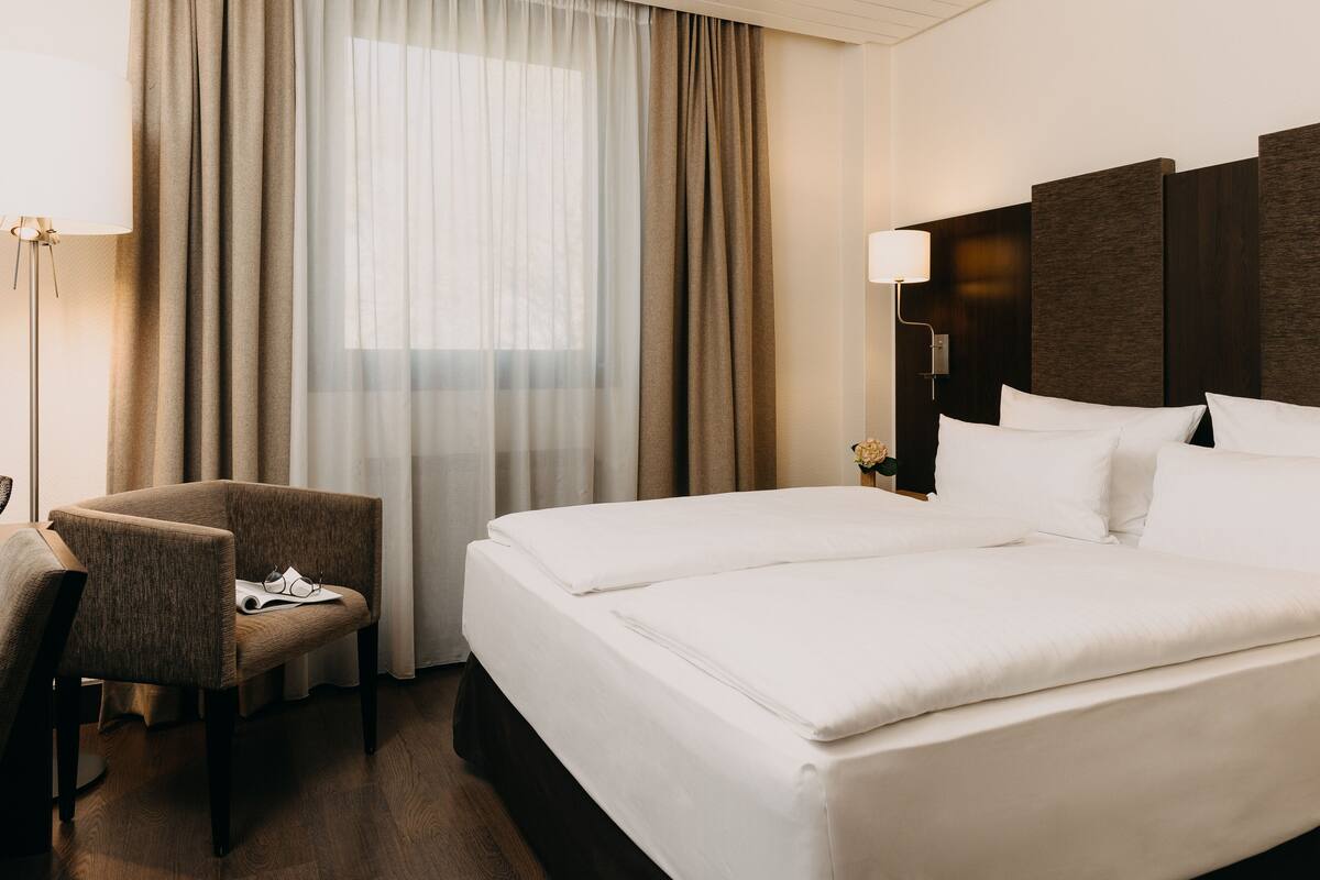 Una camera d'albergo con un letto, una sedia, una lampada e una finestra con tende.
