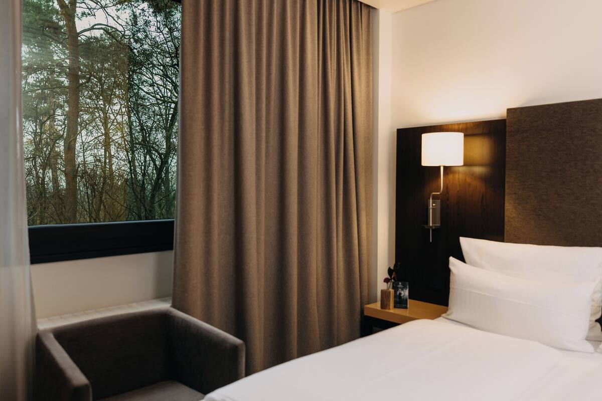 Una camera d'albergo con un letto, una sedia, un comodino con una lampada e una finestra con tende.